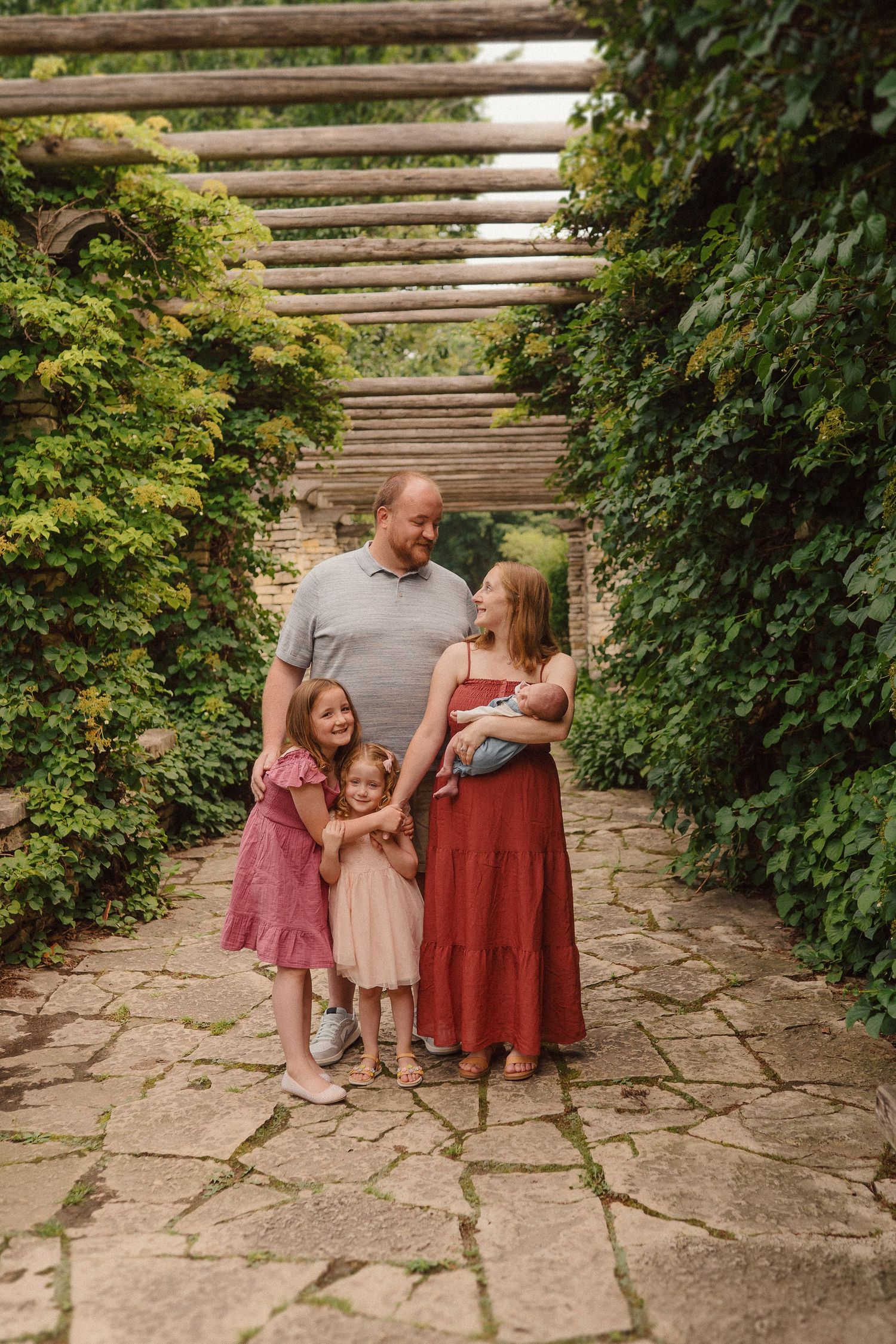 A family stands together on a stone path under a wooden pergola surrounded by lush green foliage in a garden setting.