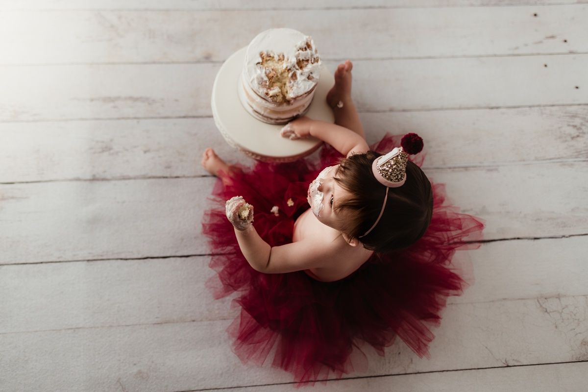 Overhead view of baby in red tutu reaching for white birthday cake during photo session.