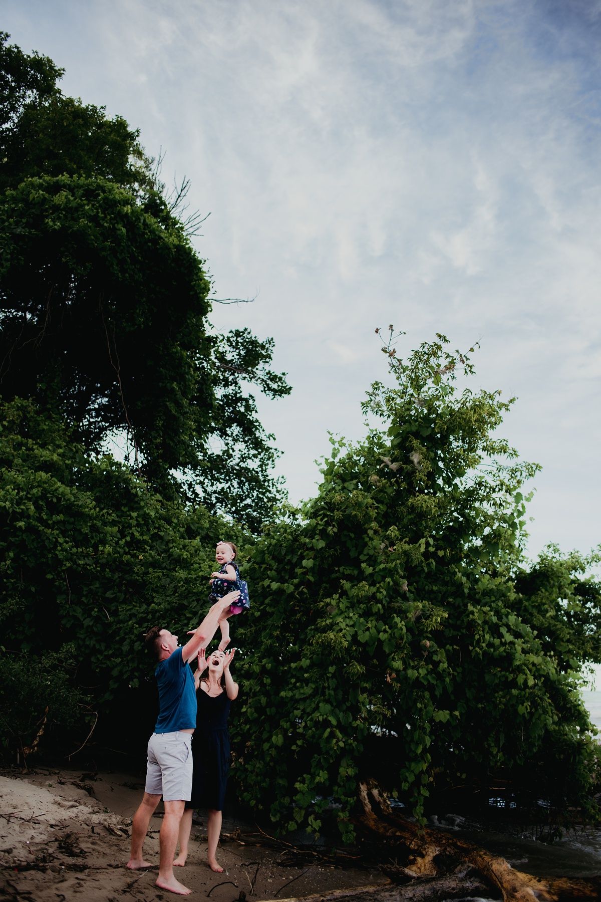 A playful family lifts a small child into the air against a backdrop of lush green trees.