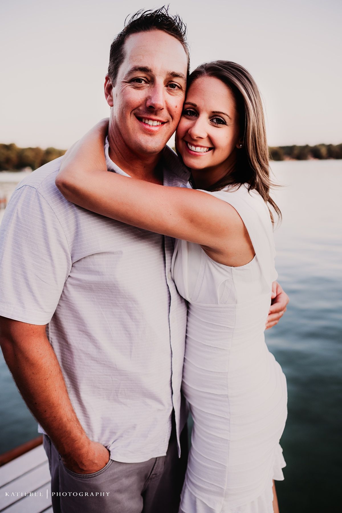 A couple in light colored clothing pose together on a boat at sunset.