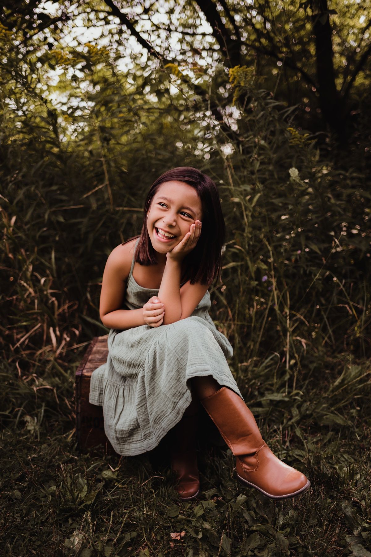 Young girl in a dress and boots sits on grass while laughing during golden hour photoshoot.