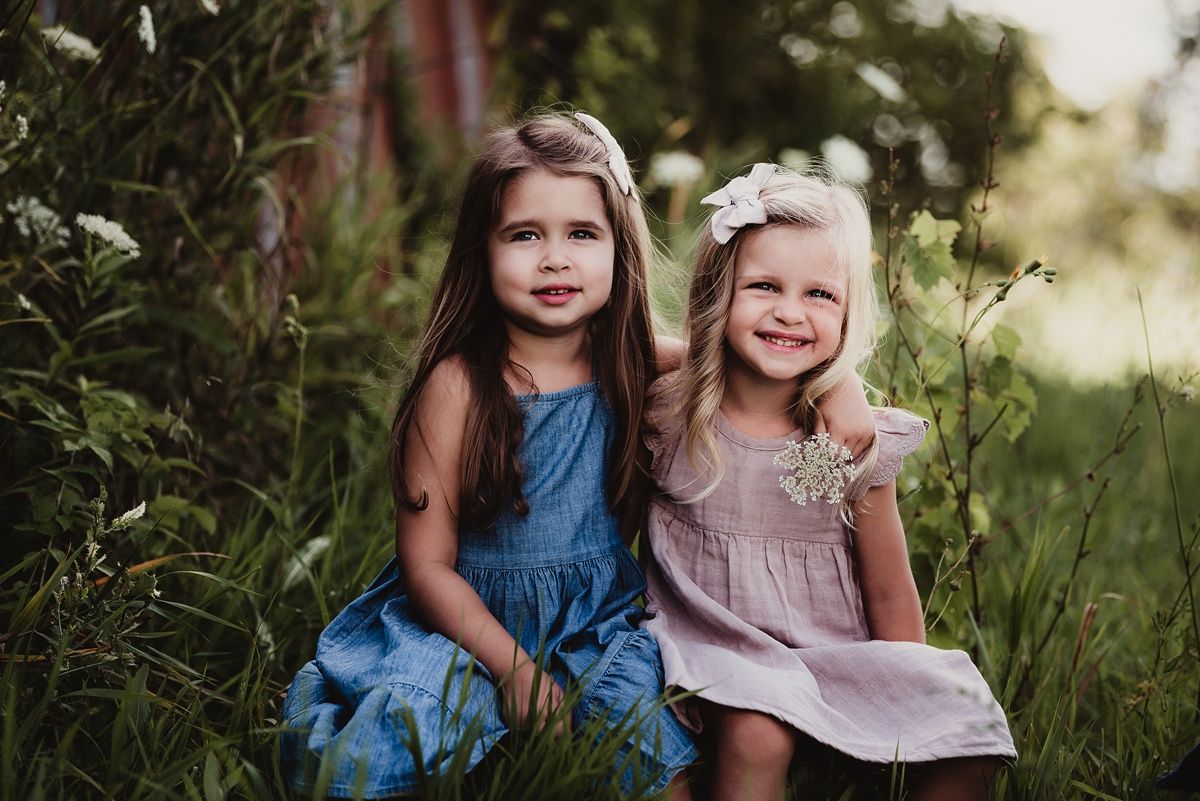 Two children in summer dresses sit together in tall grass near a barn.