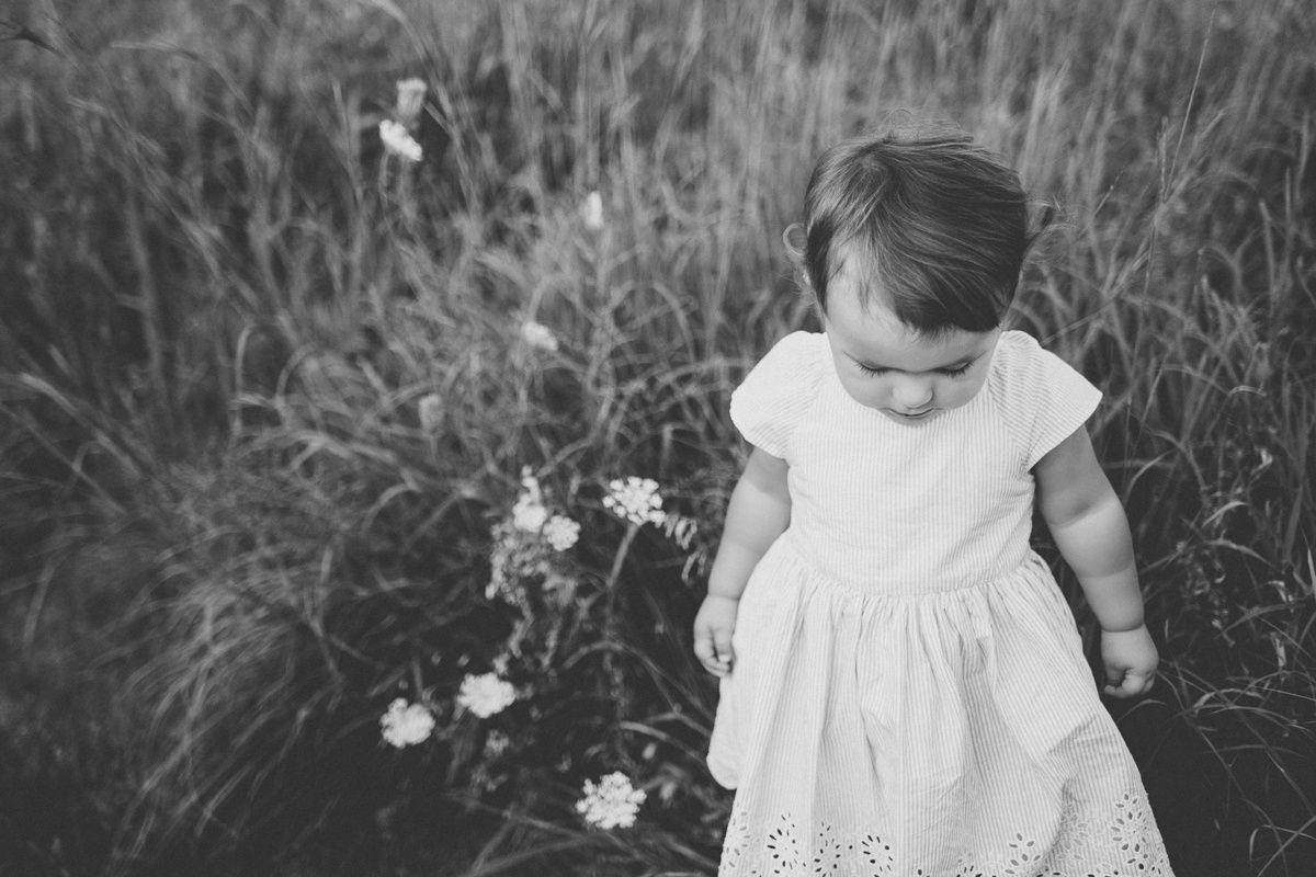 A black and white photo of a small child in a white dress standing among wildflowers.