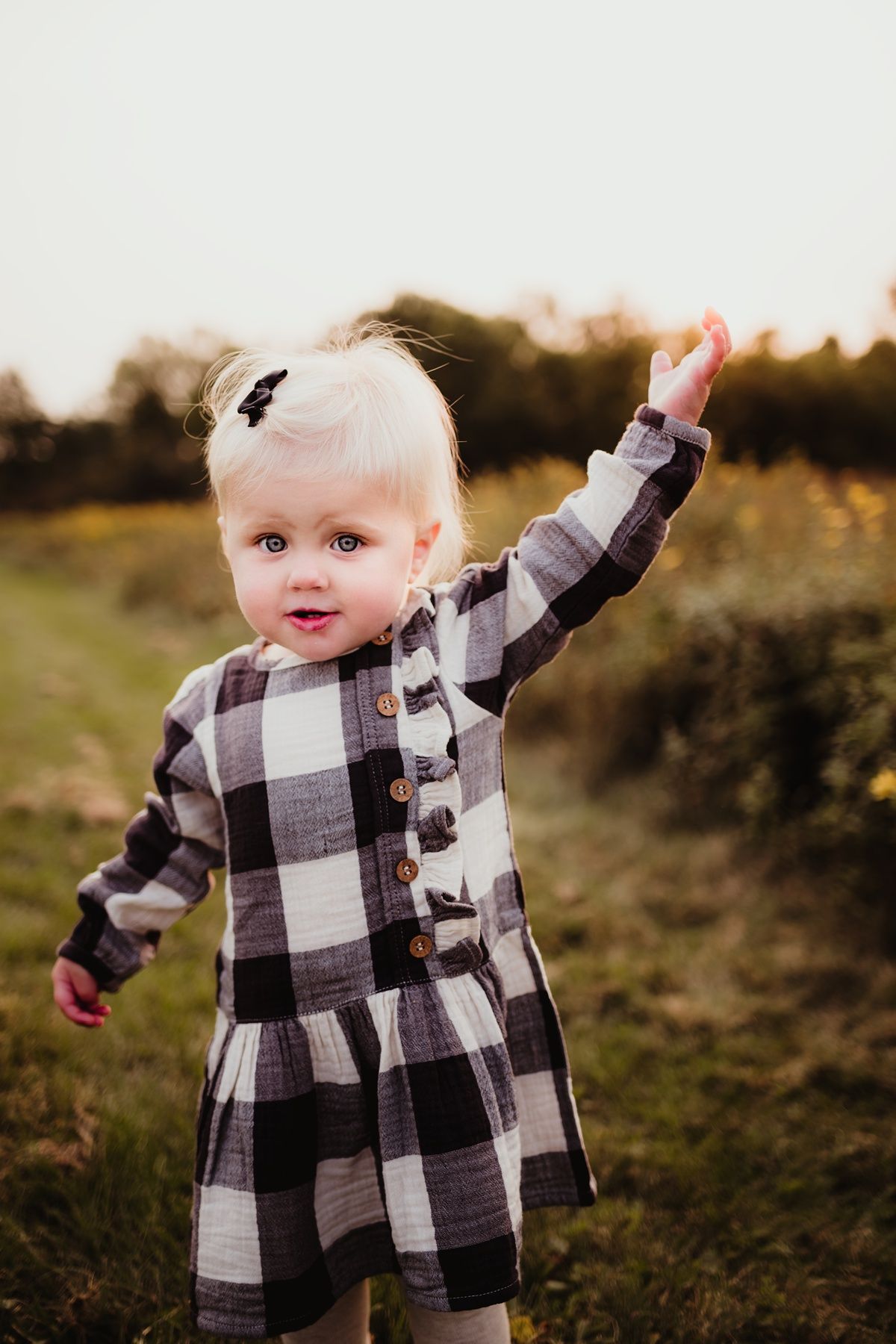 Young child in black and white plaid dress raises arm playfully against sunset backdrop in outdoor setting.