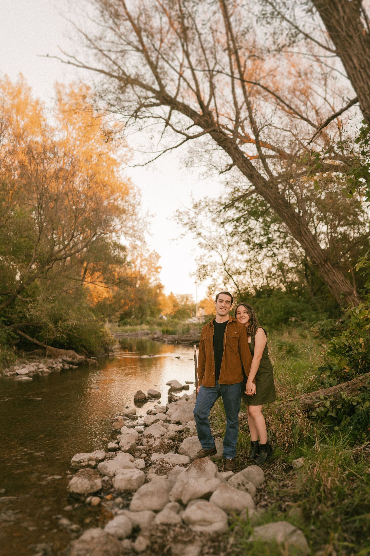 A couple stands along a rocky riverbank during golden sunset with autumn trees behind them.
