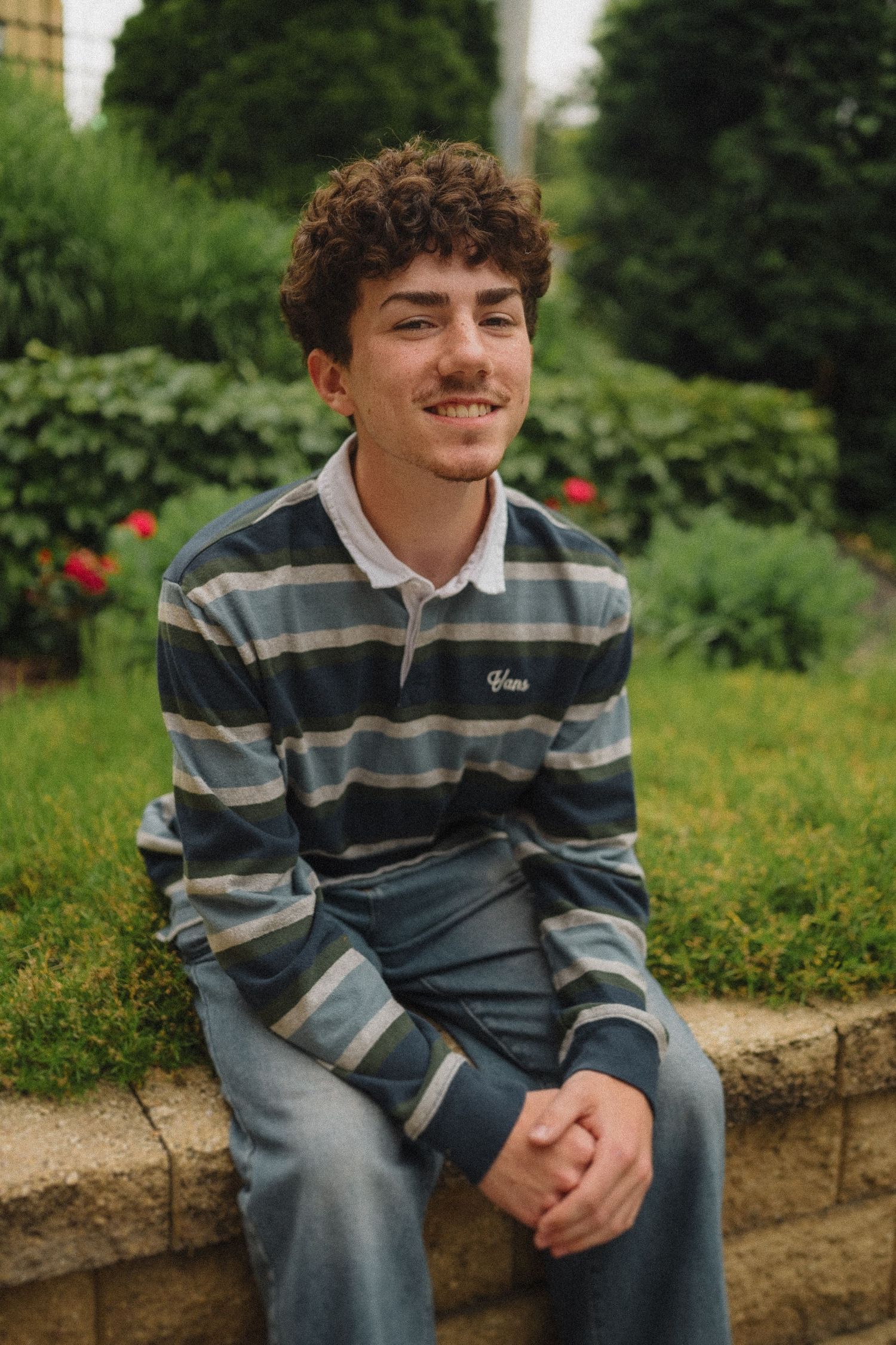 Person in a striped long-sleeve polo shirt sits smiling on a stone wall in a garden with red flowers and greenery.