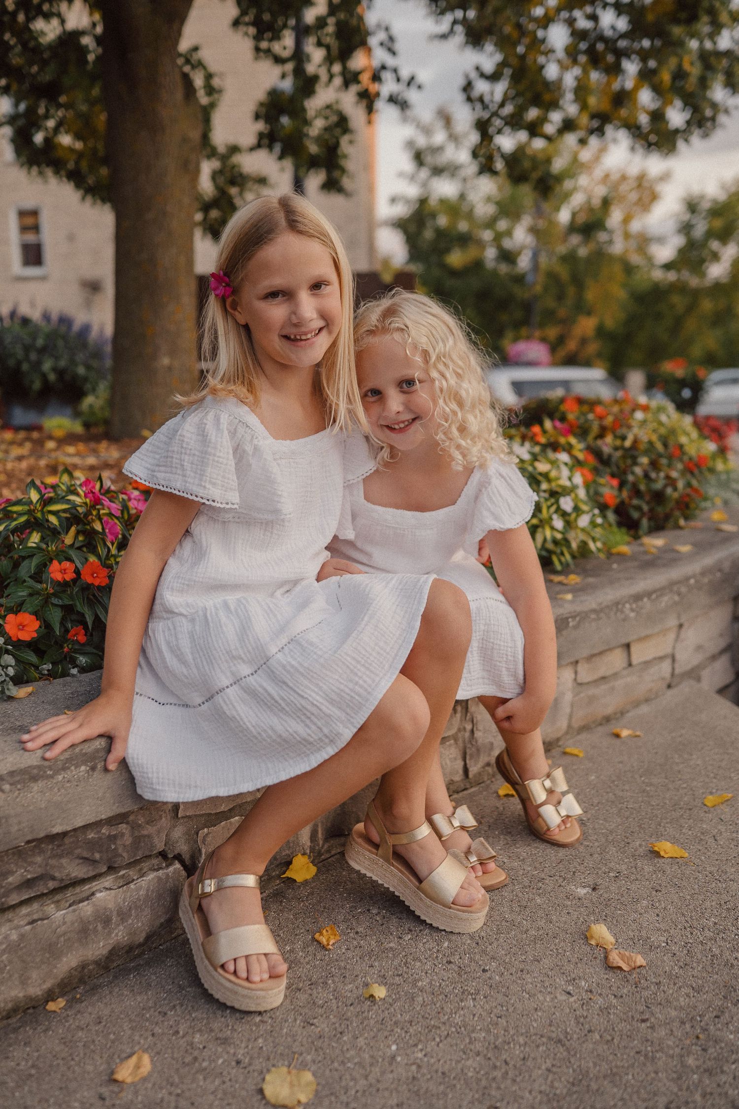 Two people in white dresses sit together on a stone curb surrounded by colorful flowers and fallen leaves.