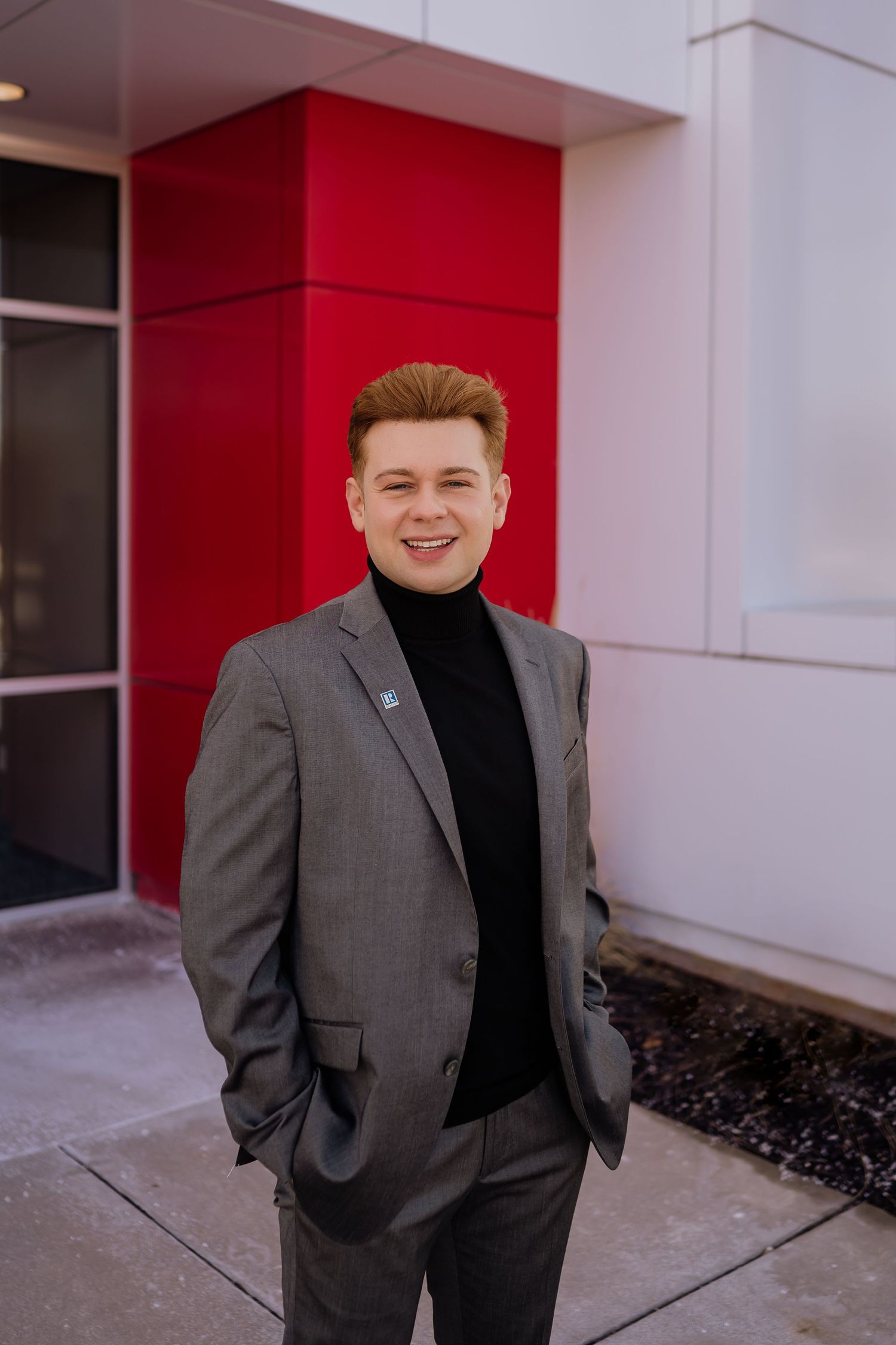 Business portrait outside modern building featuring grey suit and black turtleneck with red architectural accent.