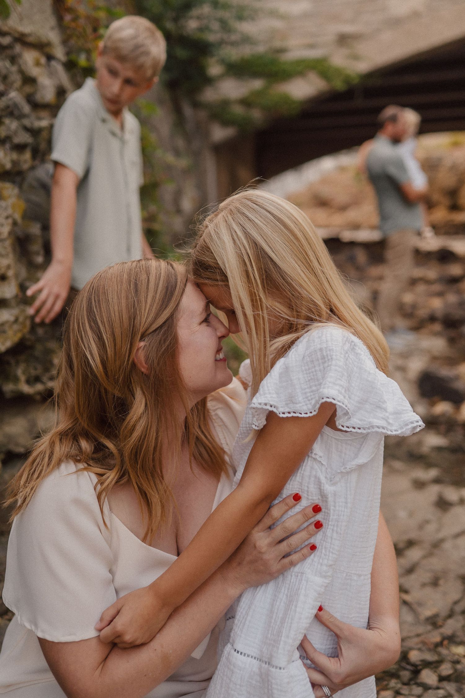 A tender family moment captured outdoors with white summer clothing against a rustic stone background.