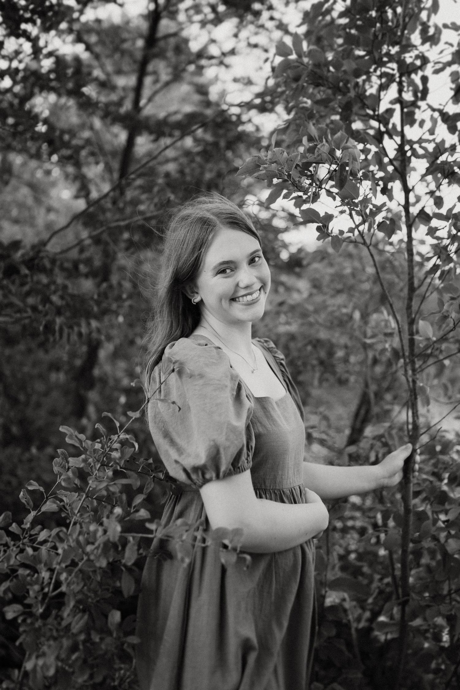 A black and white portrait showing someone in a vintage-style dress among leafy branches.