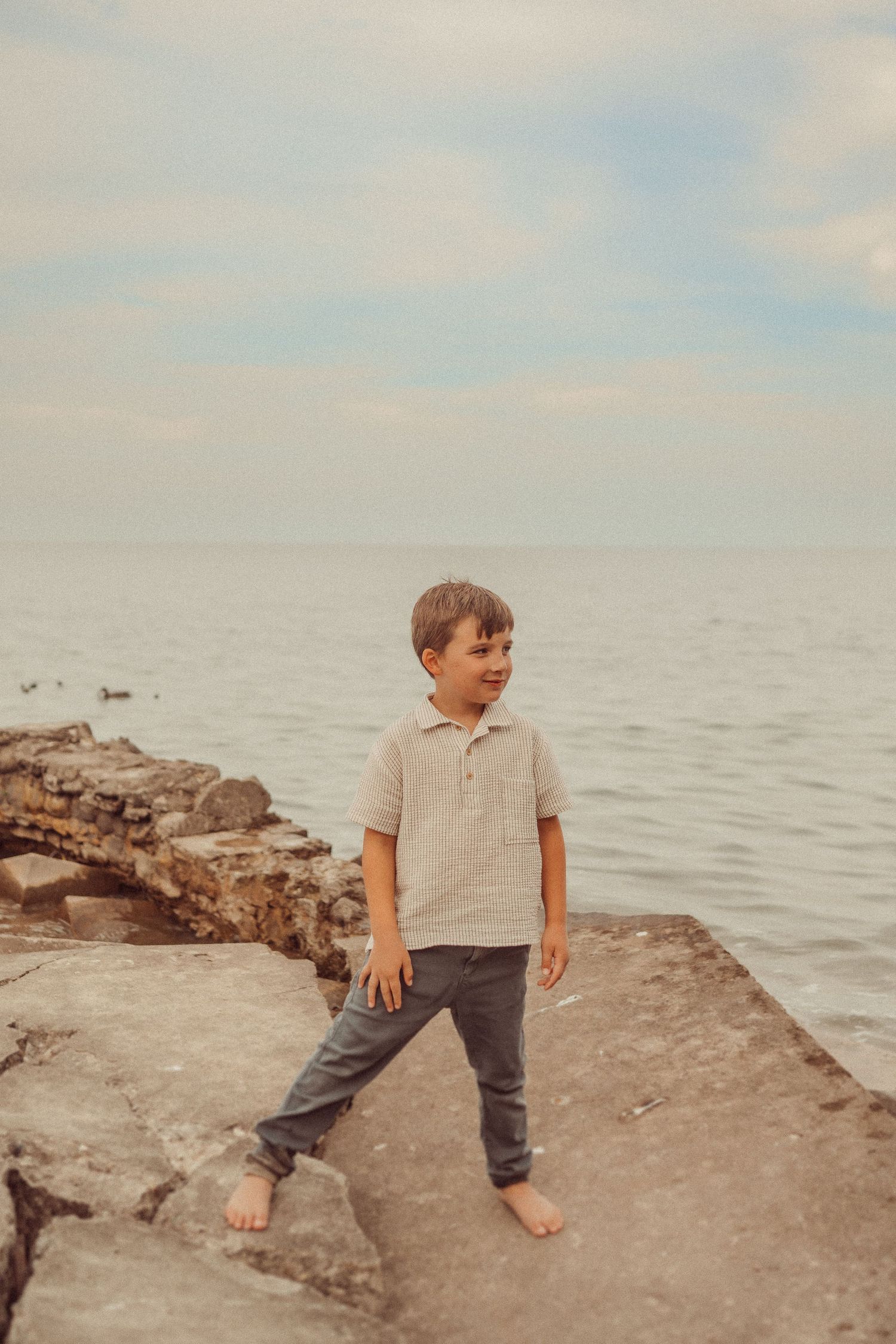 A casual portrait on rocky beach shows someone barefoot in a polo shirt and pants against the ocean backdrop.