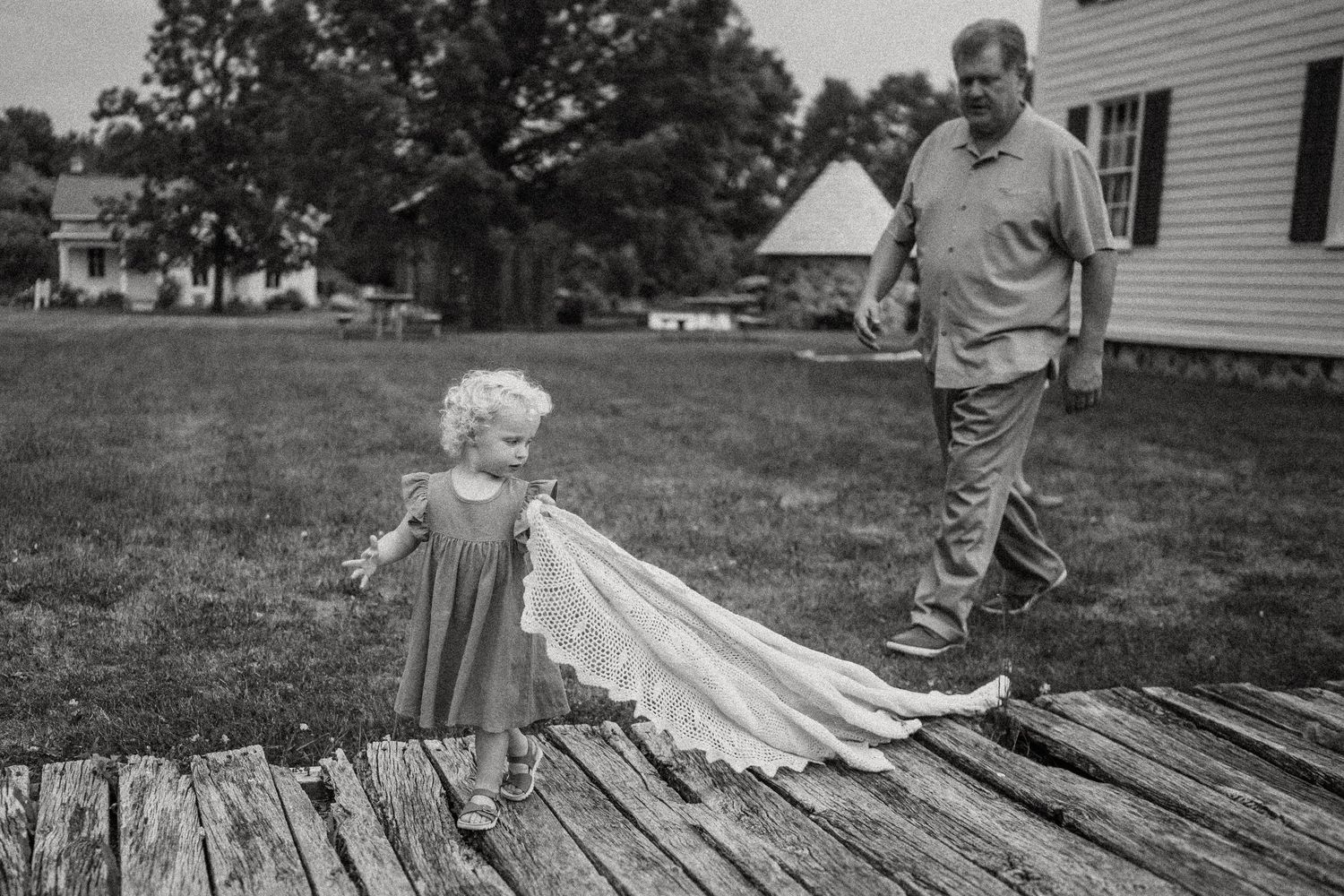 Small child walking on wooden deck with trailing dress in black and white photo.