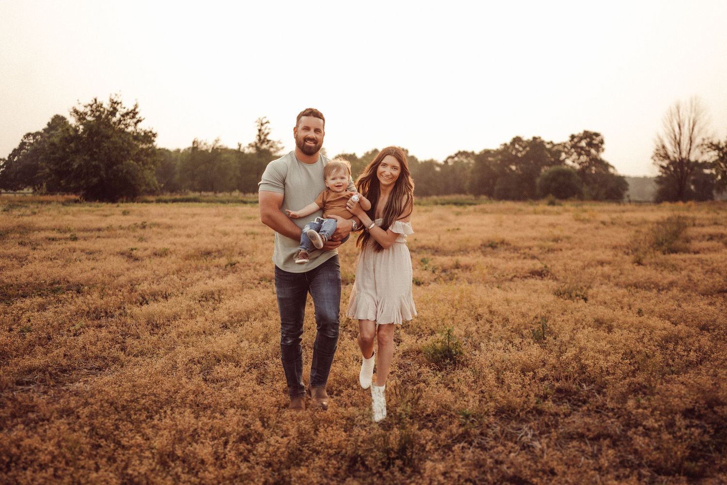 A family of three poses together in a rustic field during sunset.
