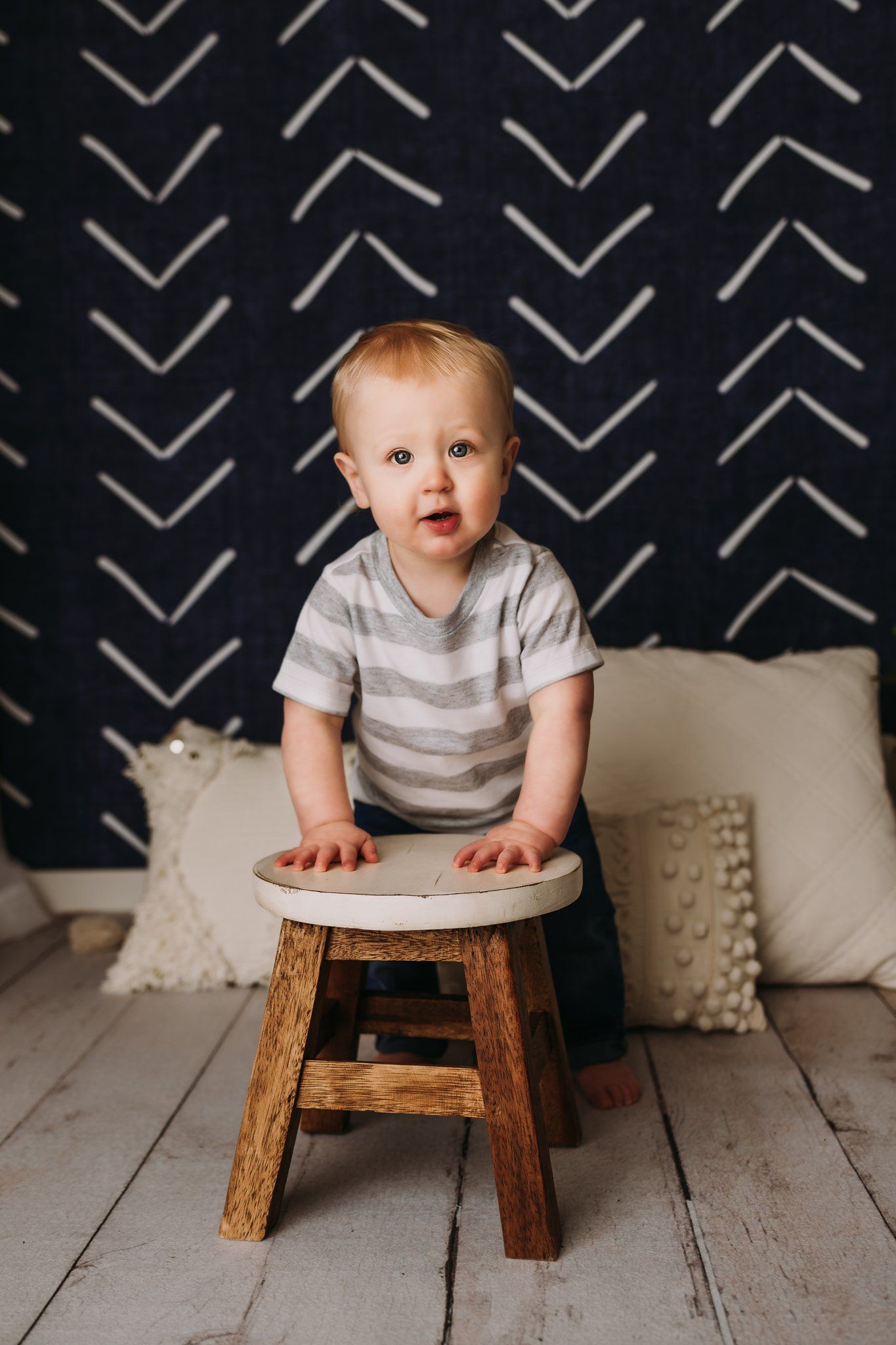 A small child stands at a wooden stool against a dark wall with chevron patterns.