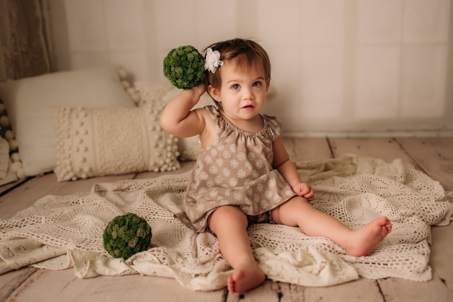 Baby in neutral dress holding green decorative ball on cream textured backdrop.