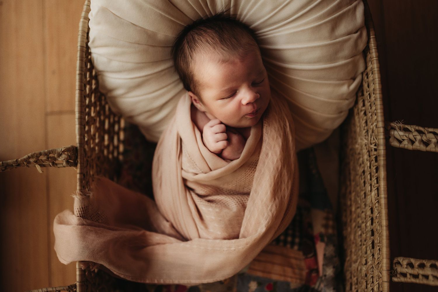 Sleeping newborn wrapped in beige fabric rests peacefully on cream pillow in wicker basket.