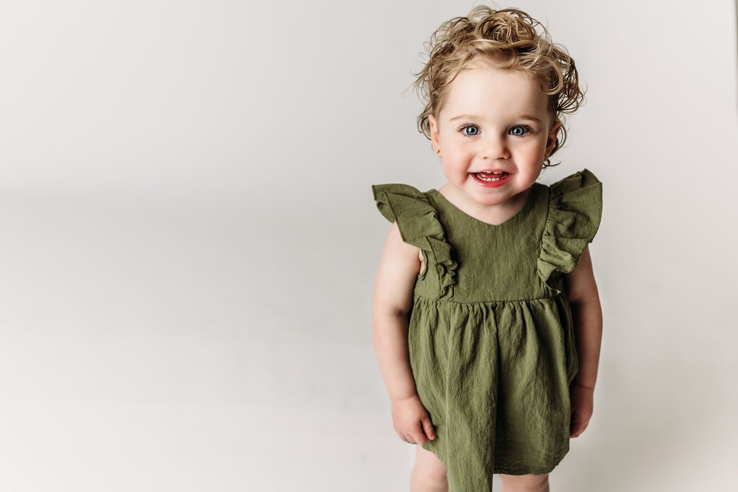 A toddler wearing a green ruffle dress smiles while standing against a white background.
