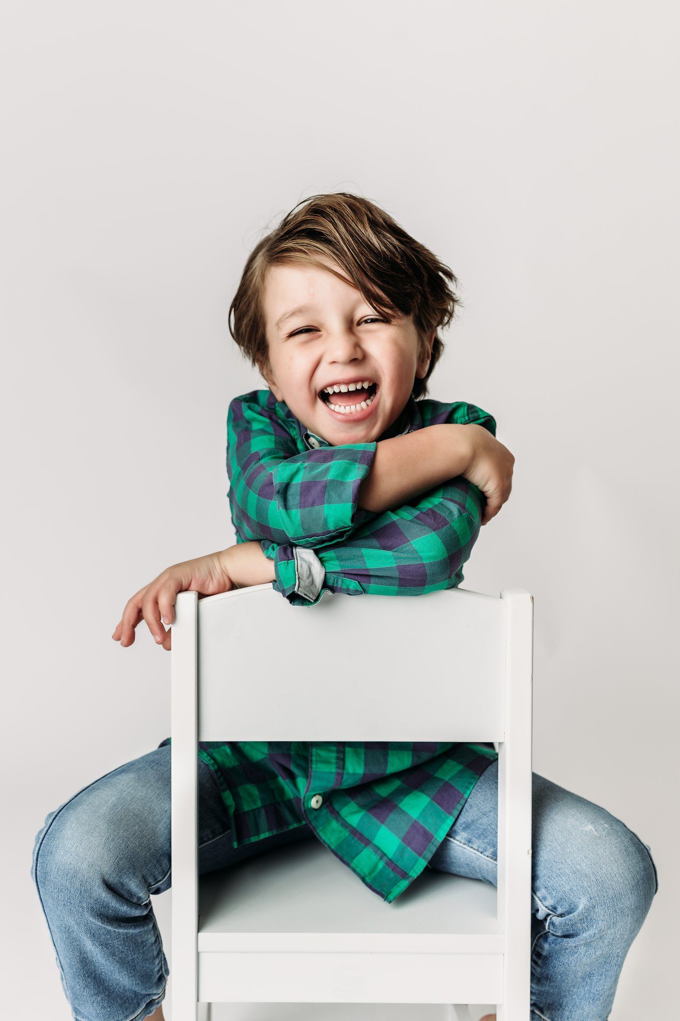 Child in plaid shirt laughs joyfully while sitting backwards on white chair.