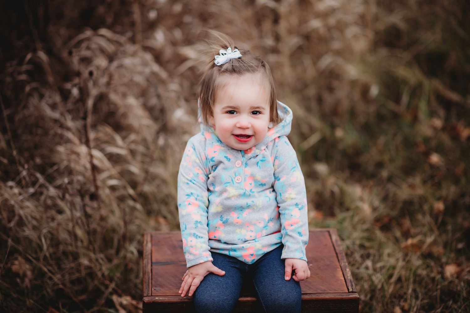 A happy toddler in floral print sitting on a wooden bench outdoors.