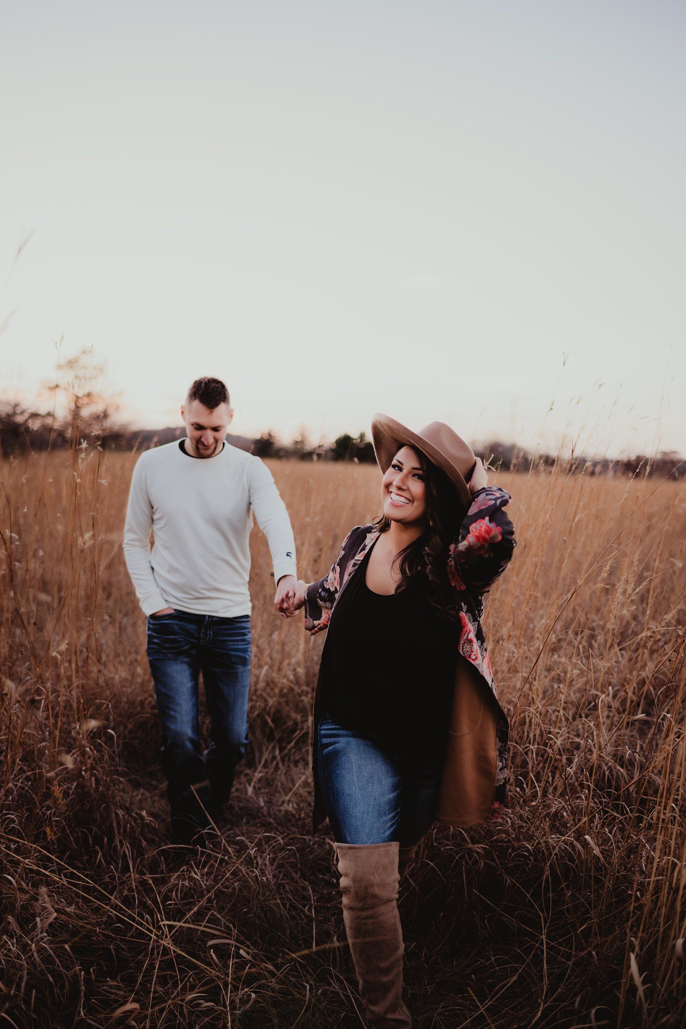 A playful couple walks through tall autumn grass with a brown hat and floral outfit.