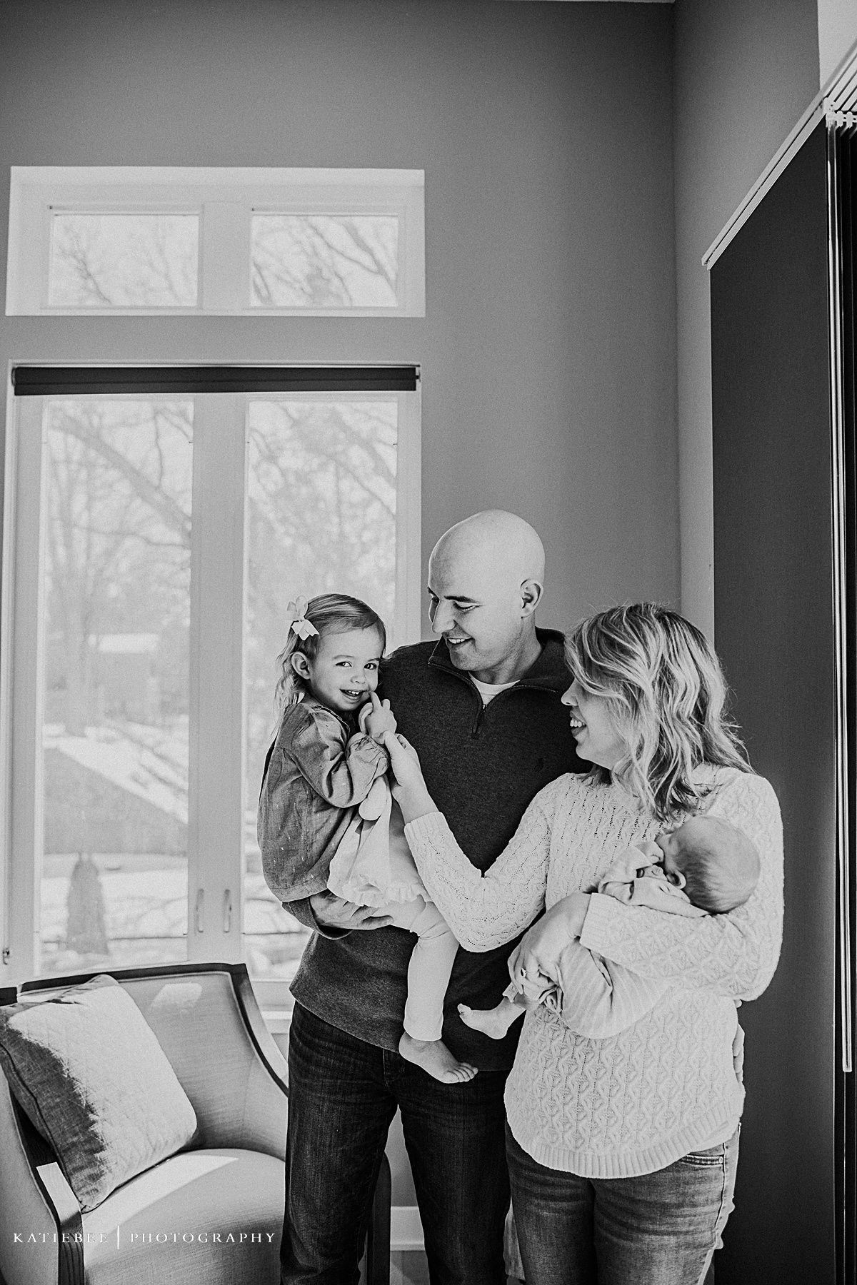 Black and white indoor family photo showing parents and children sharing intimate moments by a window.