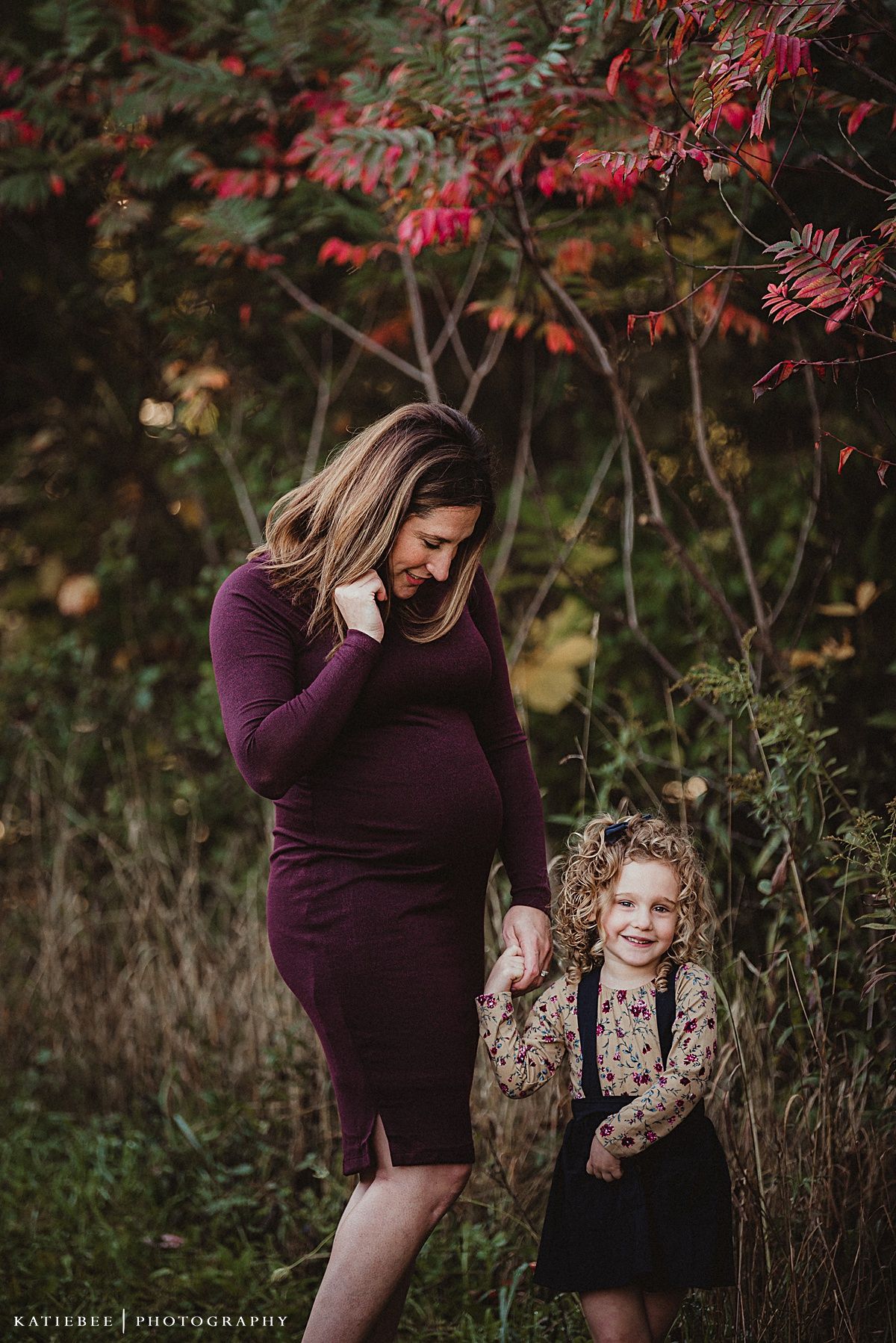 Maternity photo in autumn setting with burgundy dress and fall foliage in background.