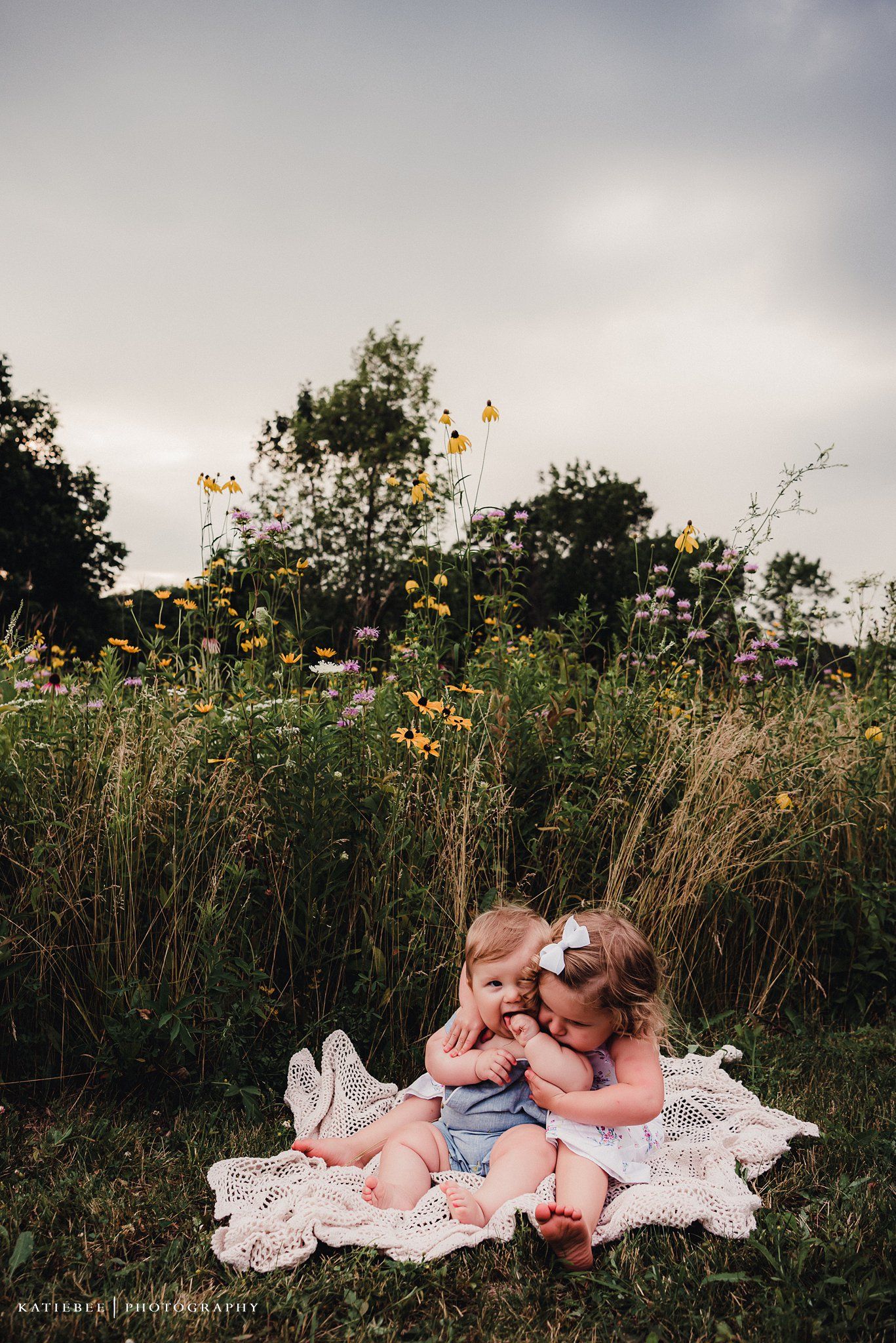 Two small children sitting together on white blanket surrounded by tall grass and yellow flowers.