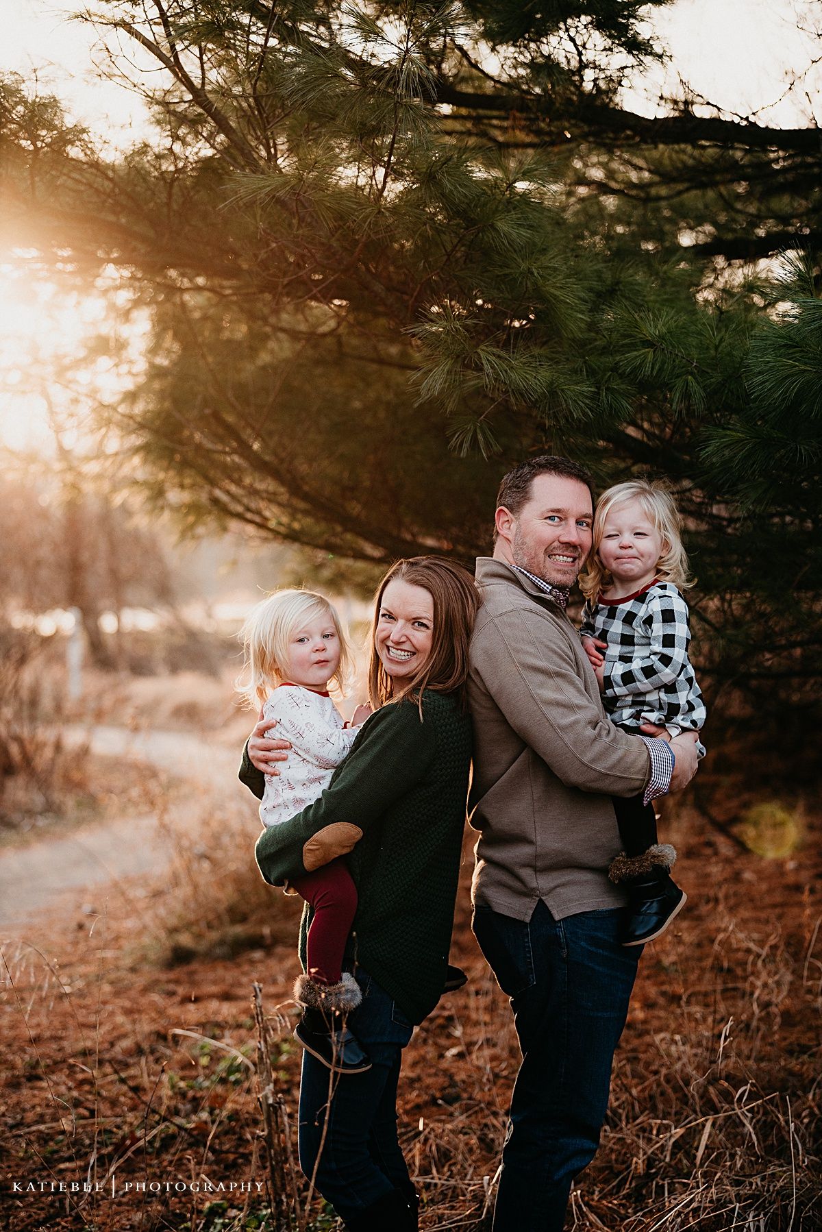 A family of four poses together for a portrait in front of pine trees during golden hour.