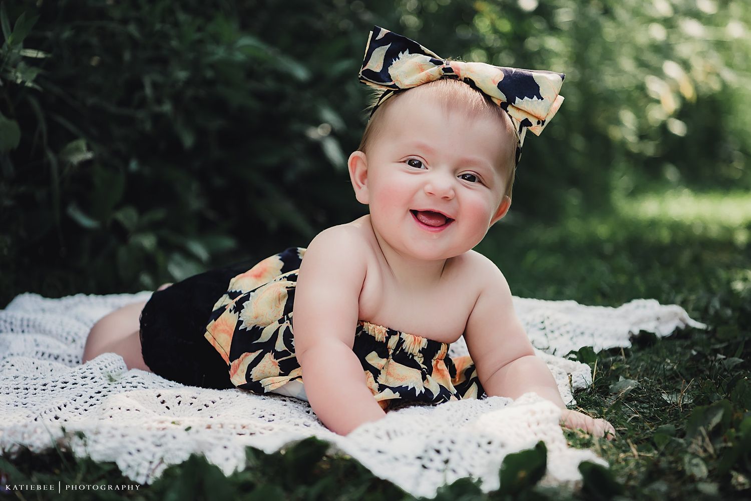 Baby wearing floral outfit laying on white lace blanket in garden setting.