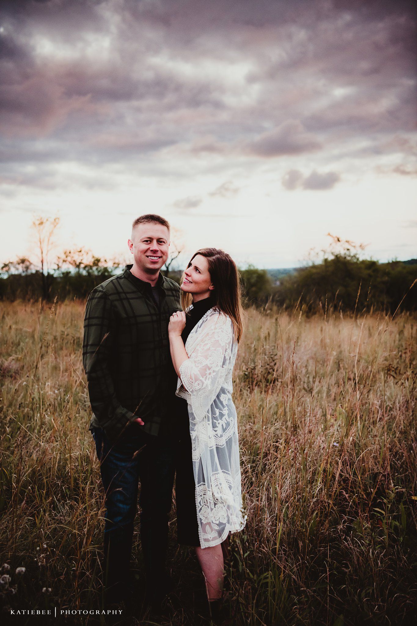 A stylish couple poses intimately in a scenic field under dramatic purple-tinted clouds.
