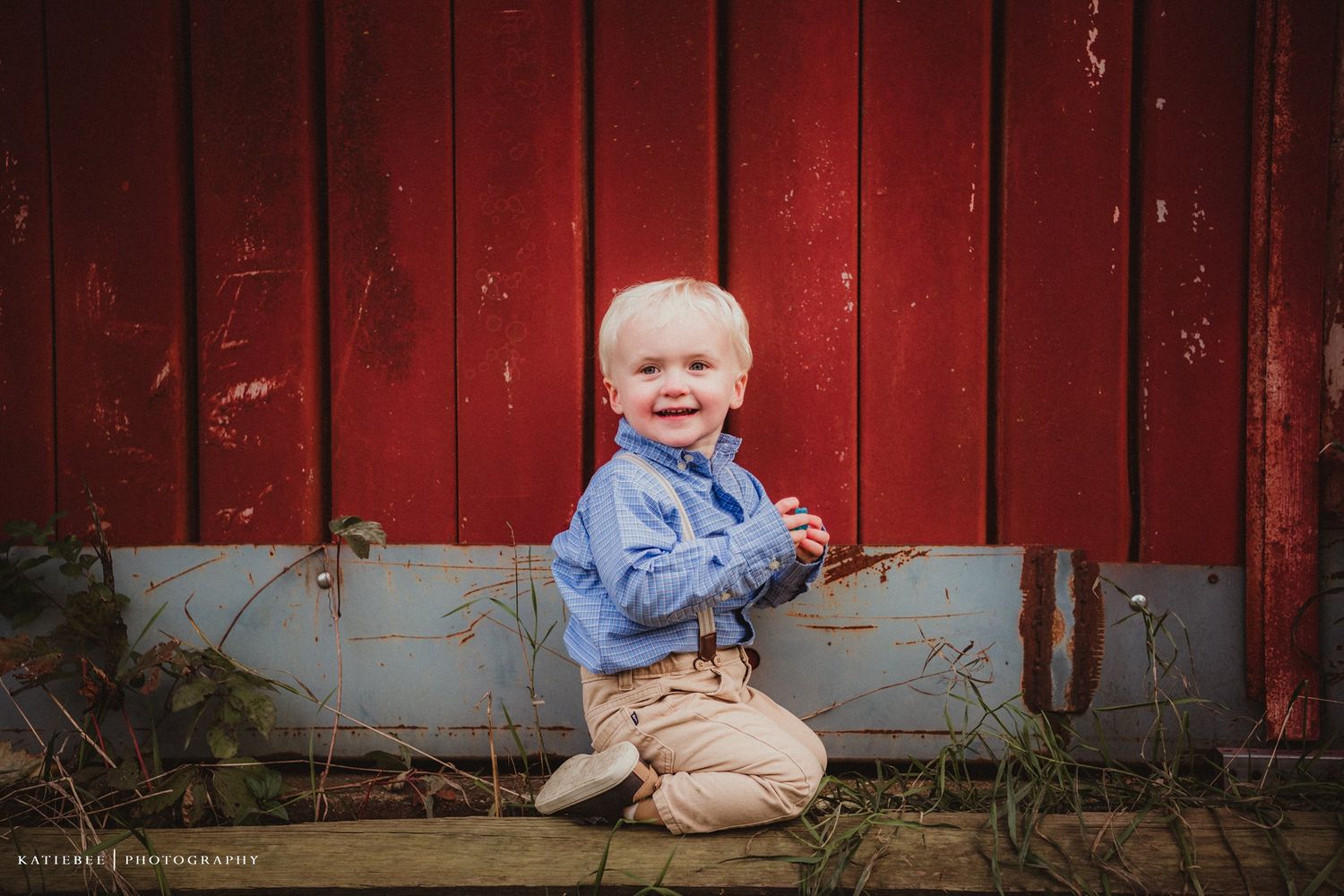 A young child sits smiling in front of a rustic red barn wall.