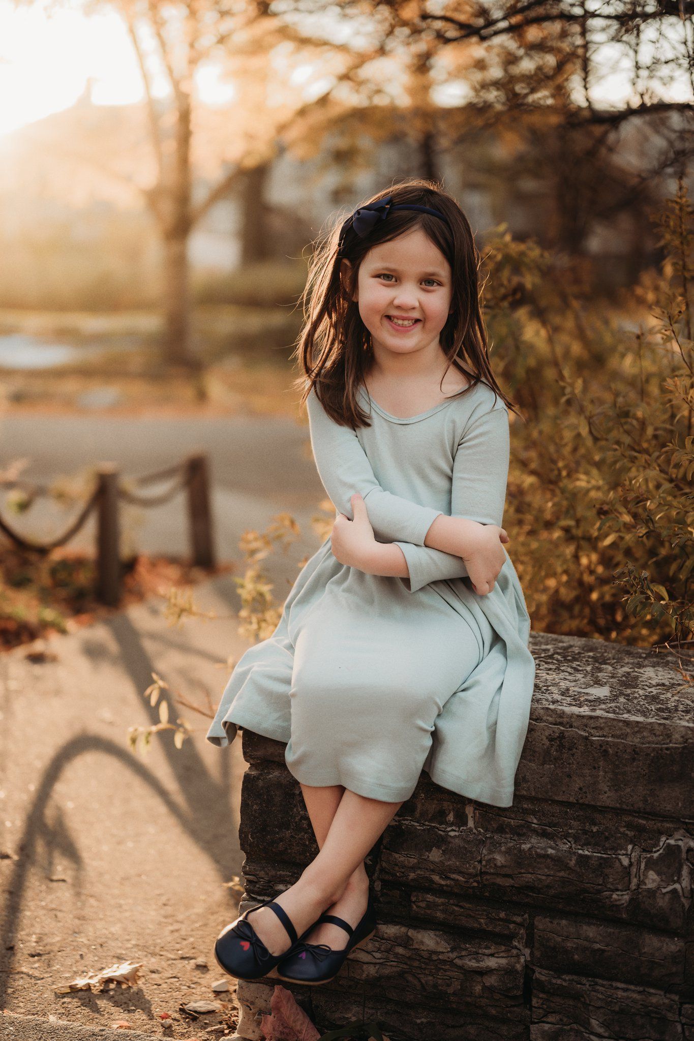 Child in blue dress sits on wooden log during golden sunset.