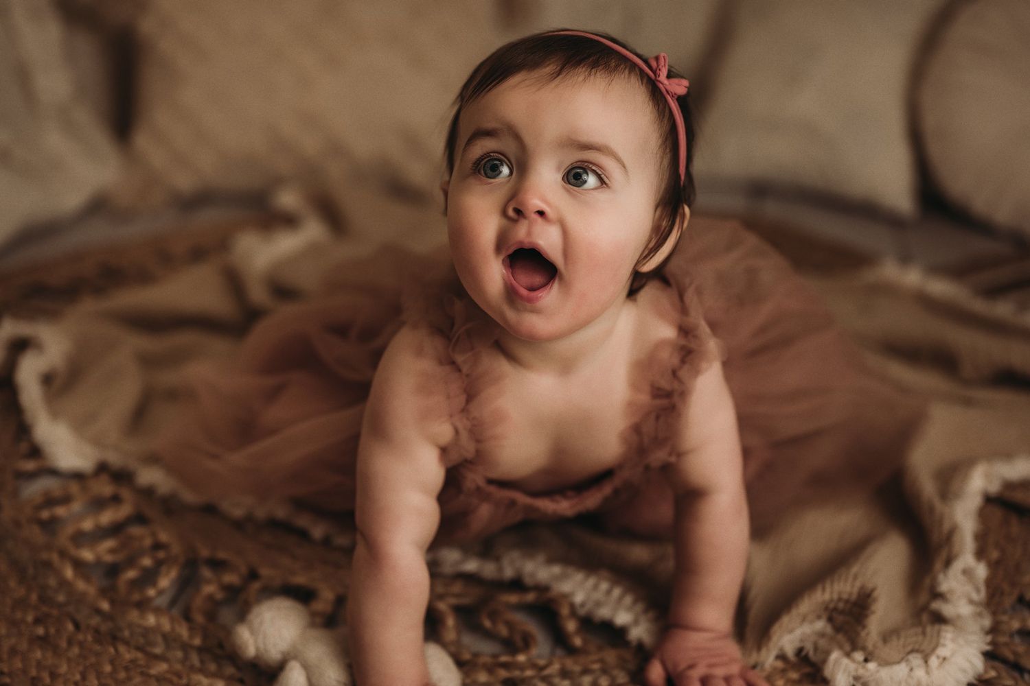 Excited baby in dusty rose dress exploring textured rug during indoor photo shoot.