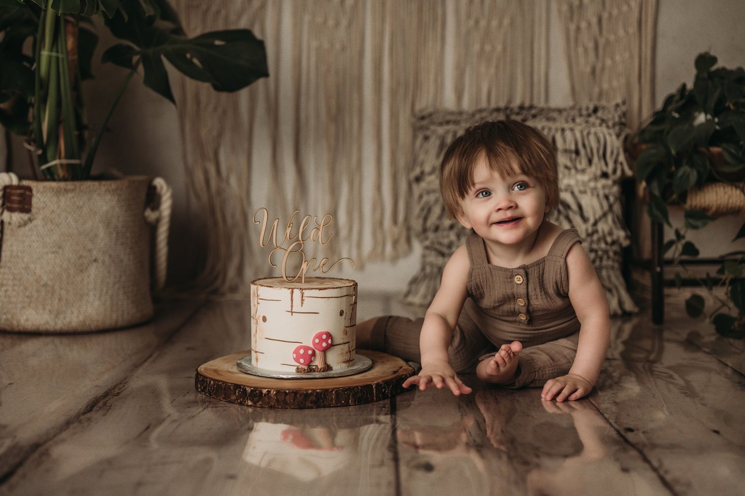 A cozy woodland-themed setup features a white cake on a wooden slice with macrame backdrop and potted plants.