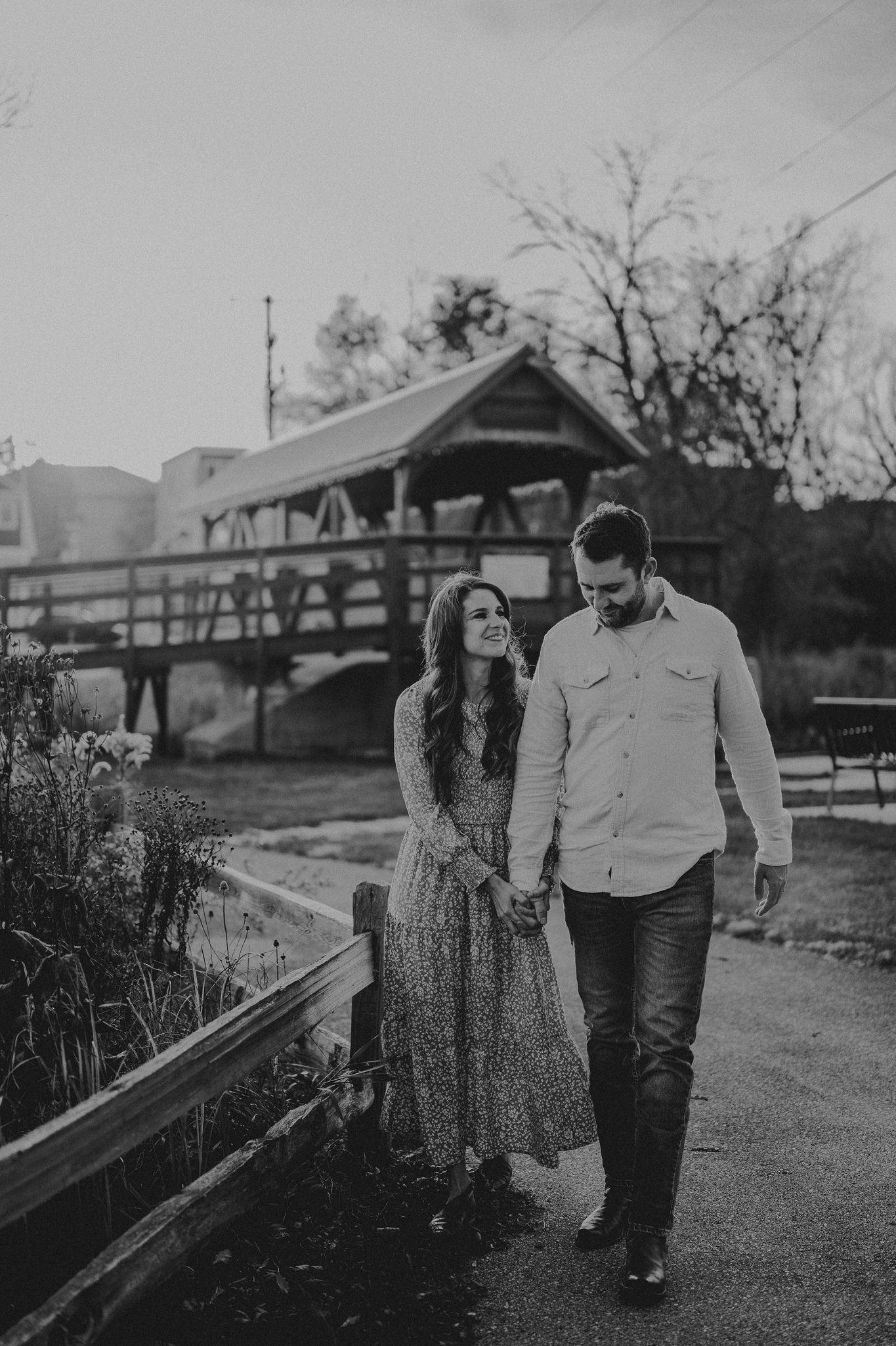Couple walking together on a path near a wooden bridge in black and white photography.