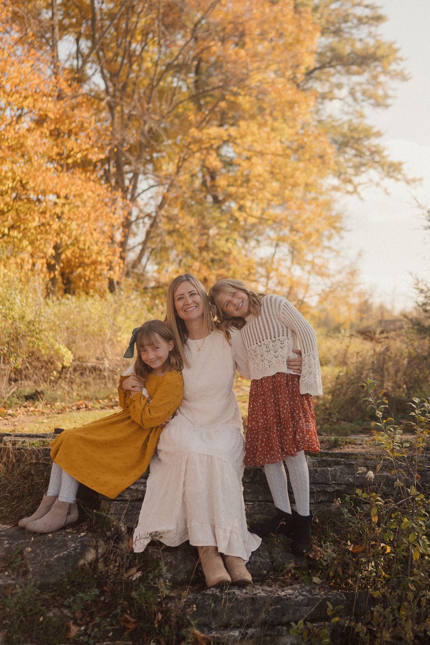 Three people sitting together on a stone wall surrounded by golden fall foliage.