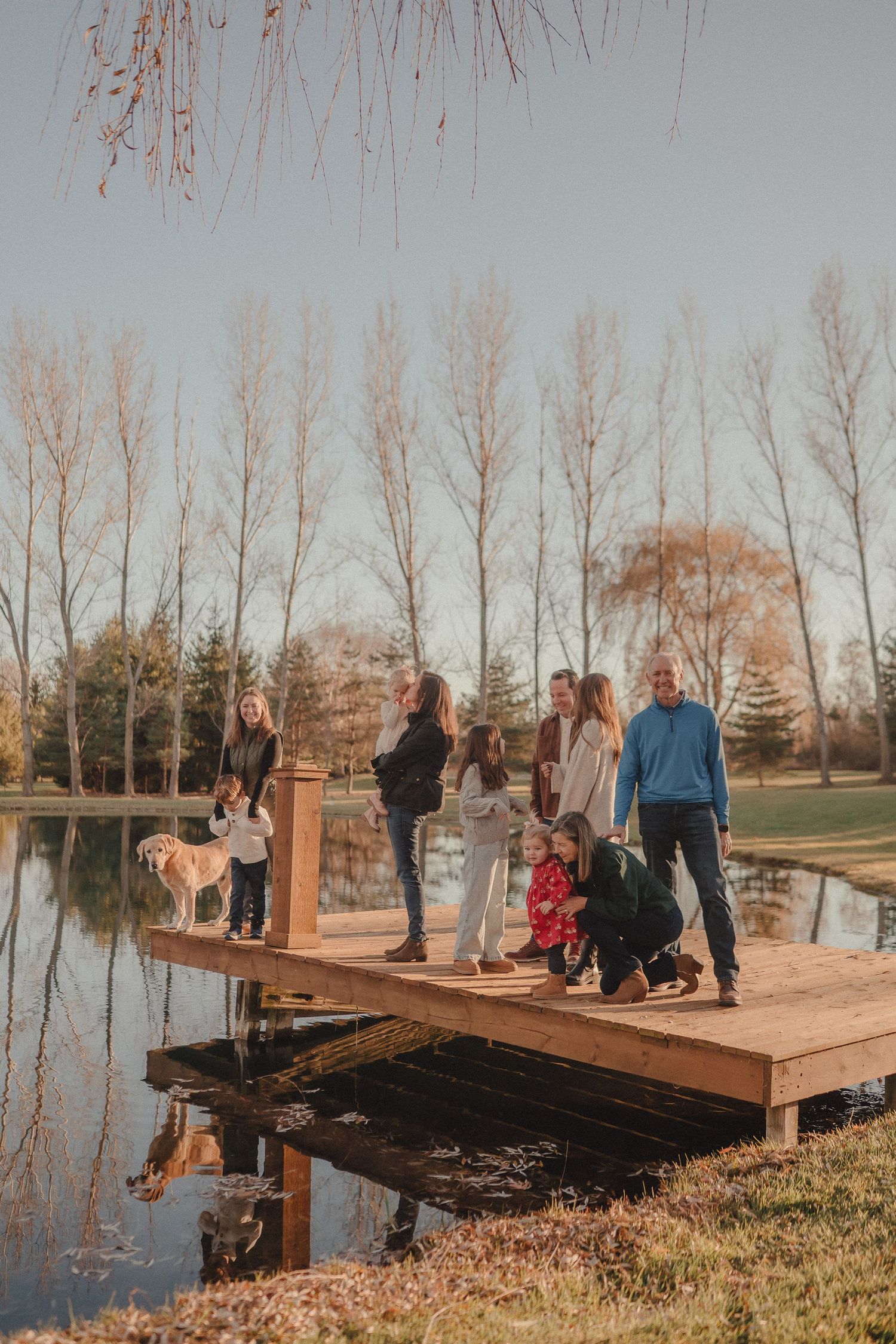 A wooden dock extends over a calm pond with bare trees reflecting in the water during autumn.