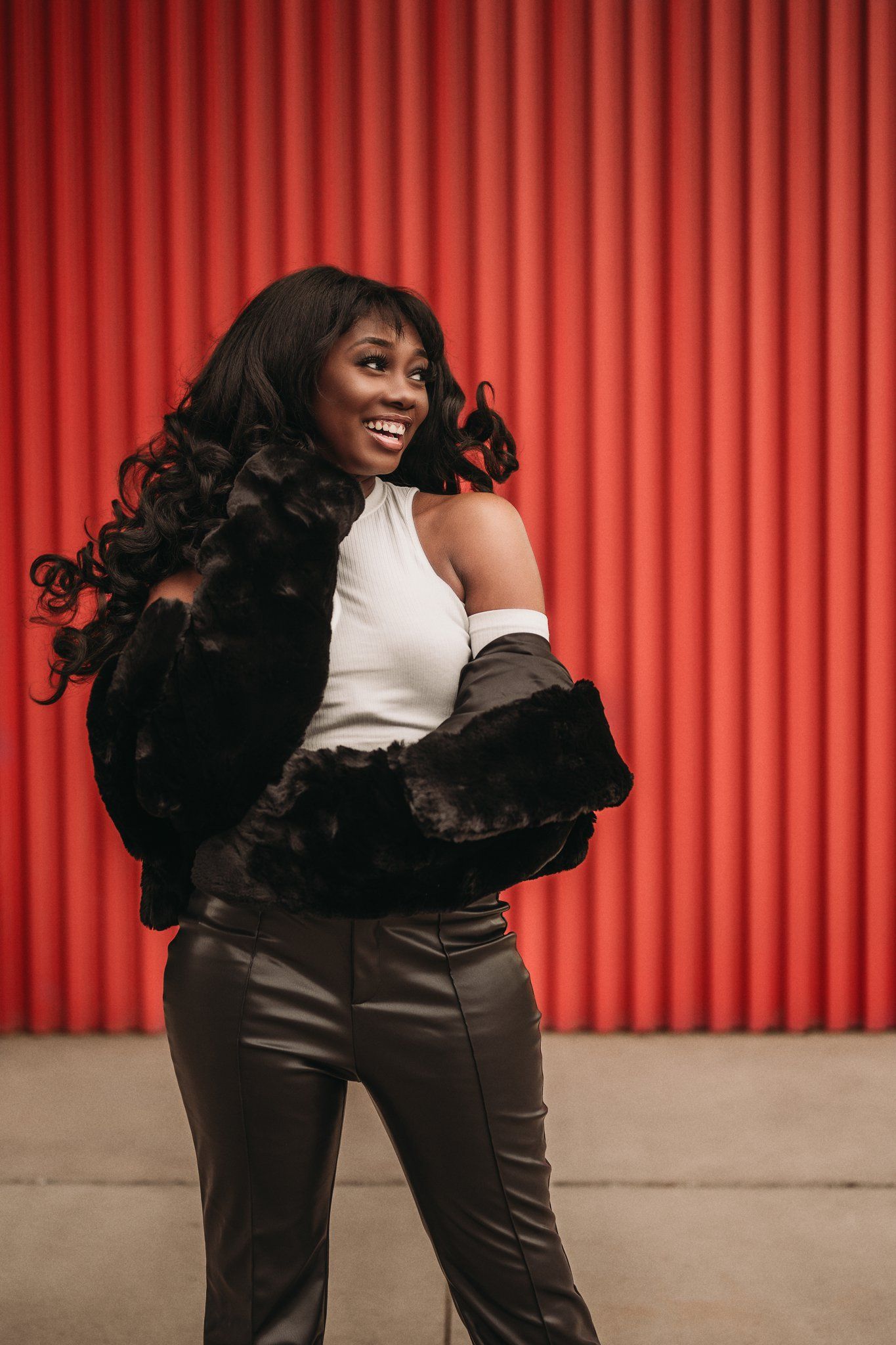 A joyful fashion model poses in a white halter top and dark pants against a red wall.