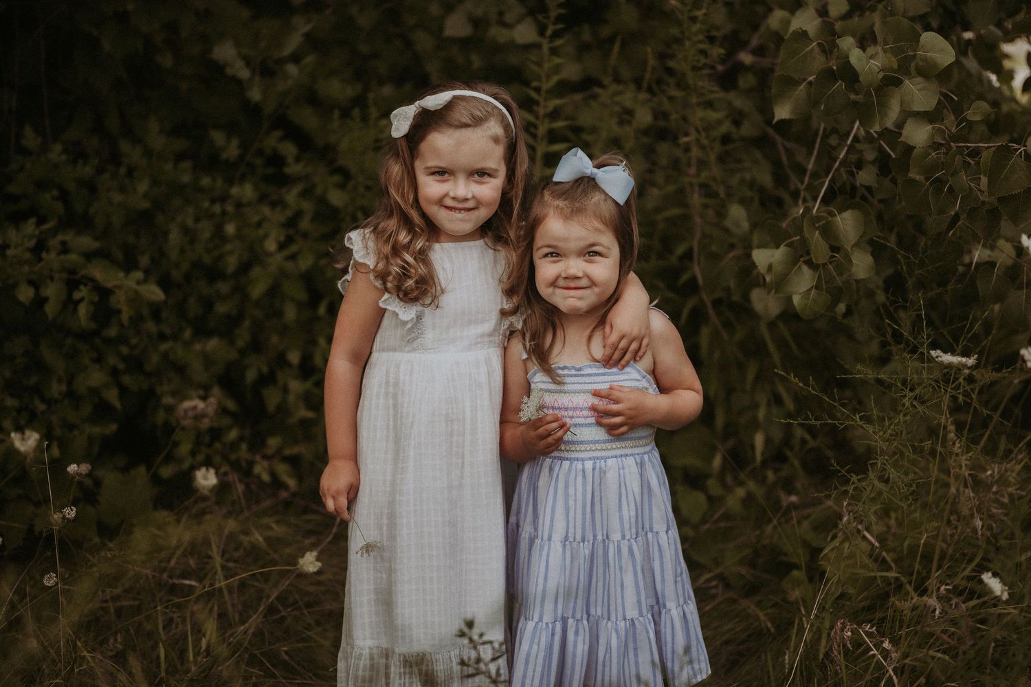 Two young children in flowy dresses stand together against a natural backdrop of green foliage.