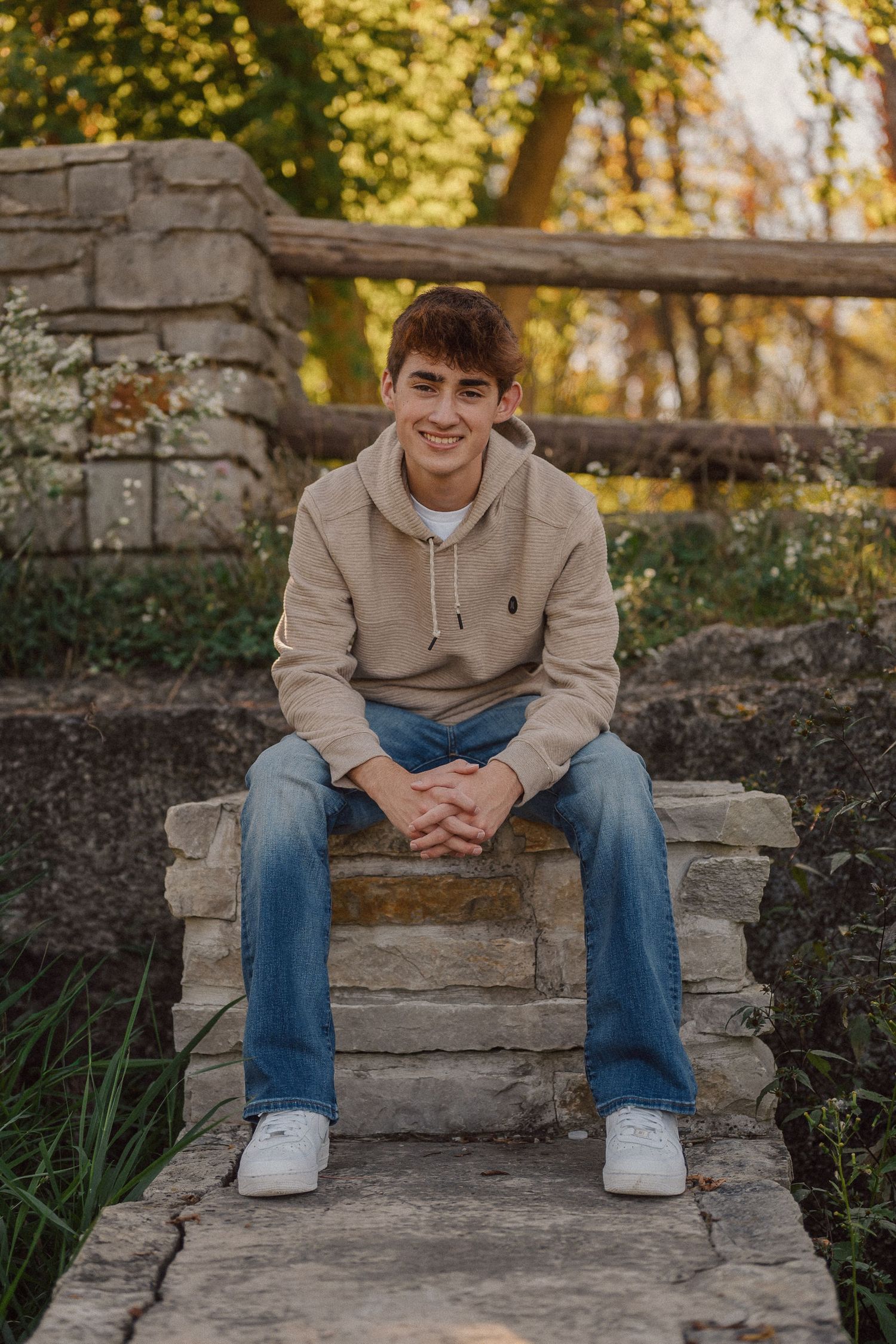 Person in beige hoodie sits on stone steps with autumn scenery and trees in background.