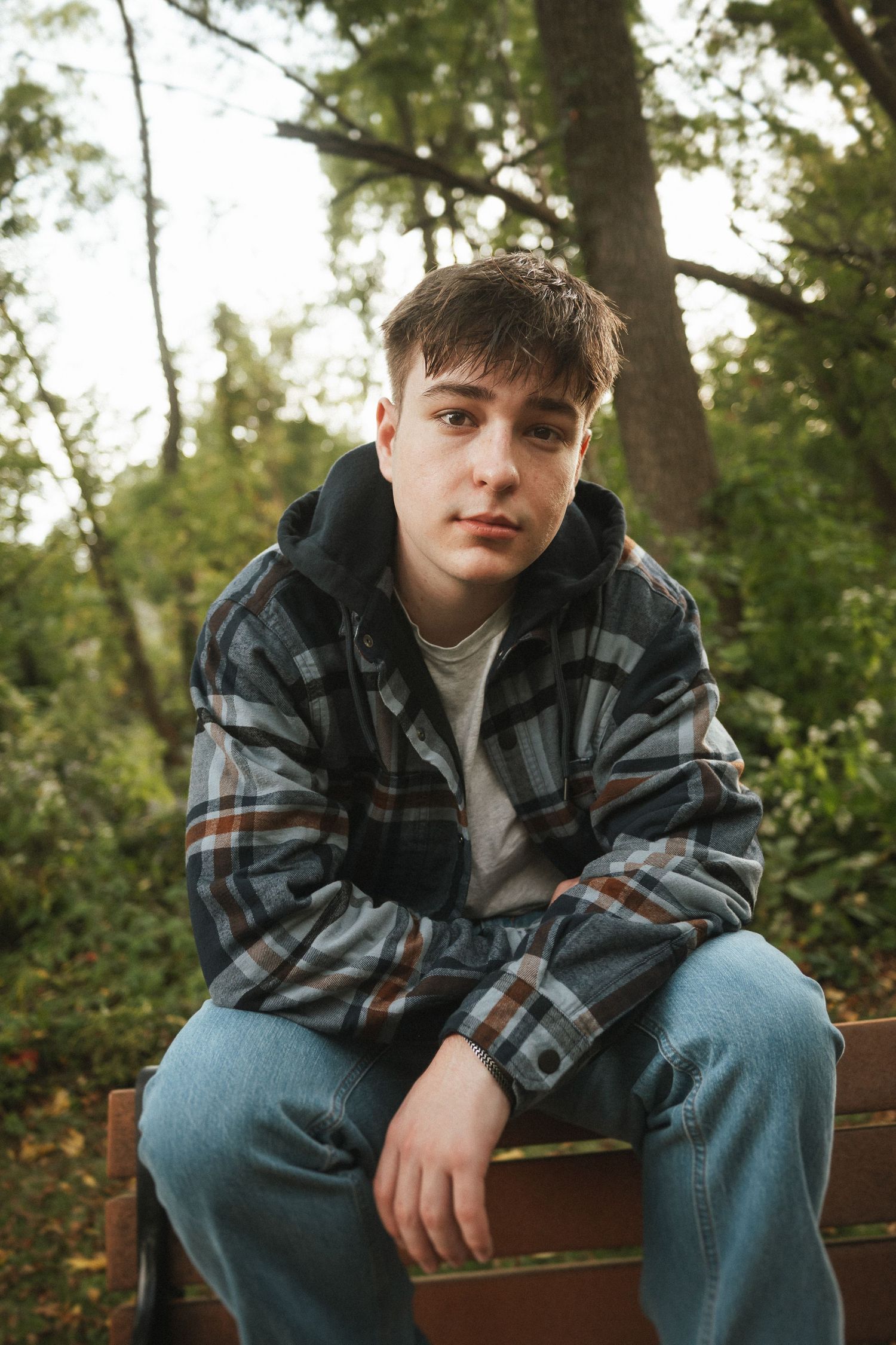 Person sitting casually on a wooden bench in a wooded area wearing a plaid jacket and jeans.