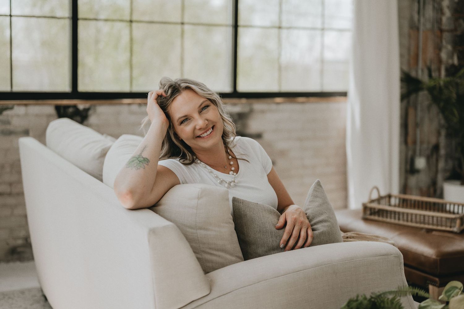 Relaxed woman lounges on white couch in stylish sunlit room with industrial windows.