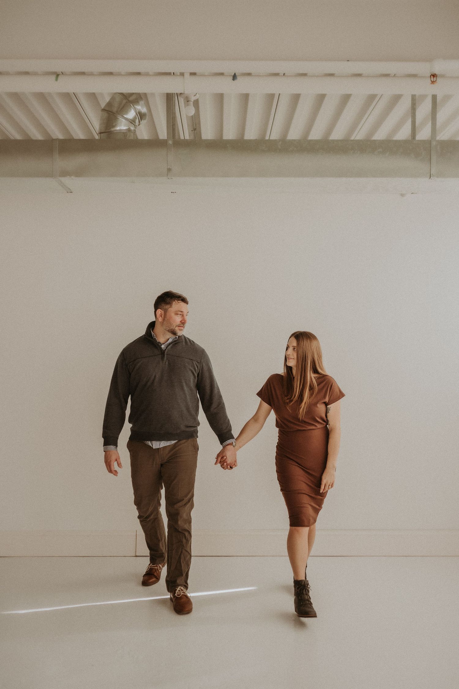 A couple walks hand in hand in a minimal white studio space with industrial ceiling.