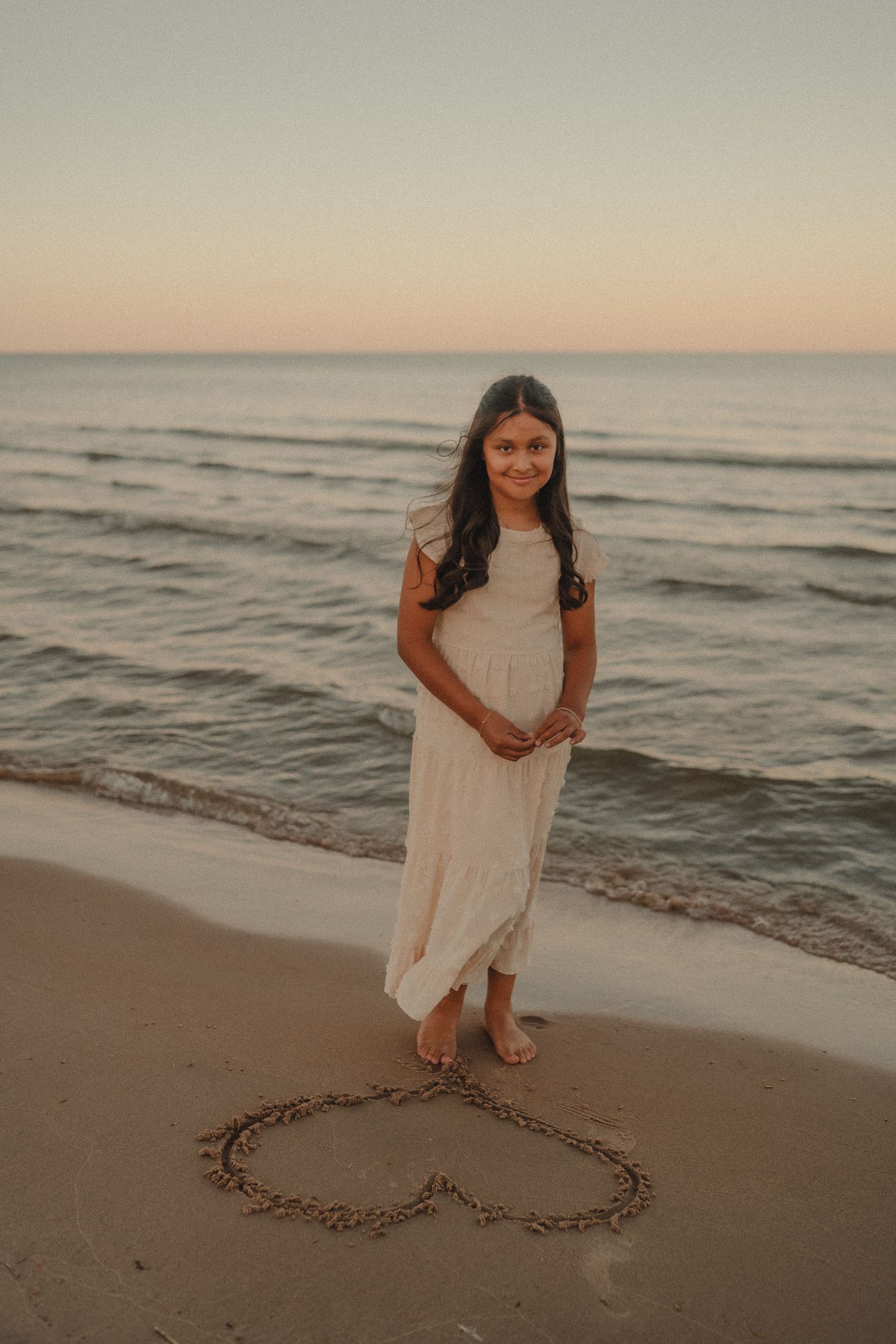 Young girl in white dress standing barefoot by ocean with heart drawn in sand at sunset.