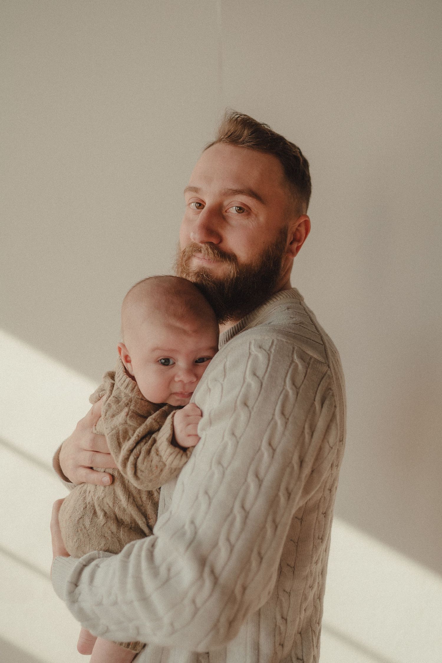 Parent and baby in matching beige cable knit sweaters share a loving embrace in natural window light.