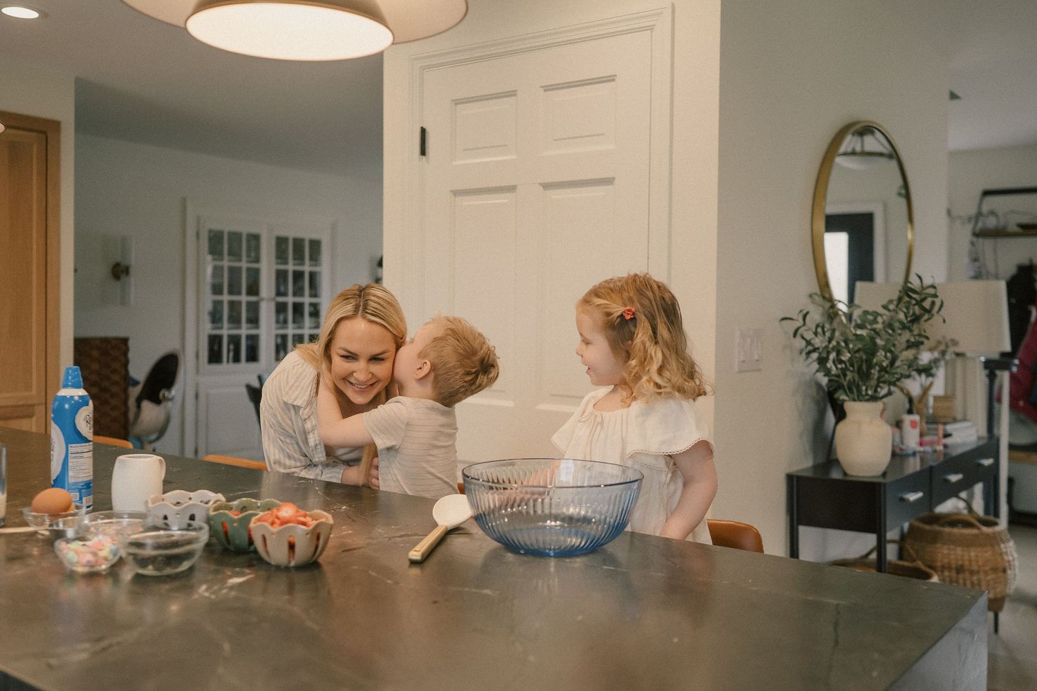 A mother and two young children laugh together while baking at a kitchen island in a bright, modern home.