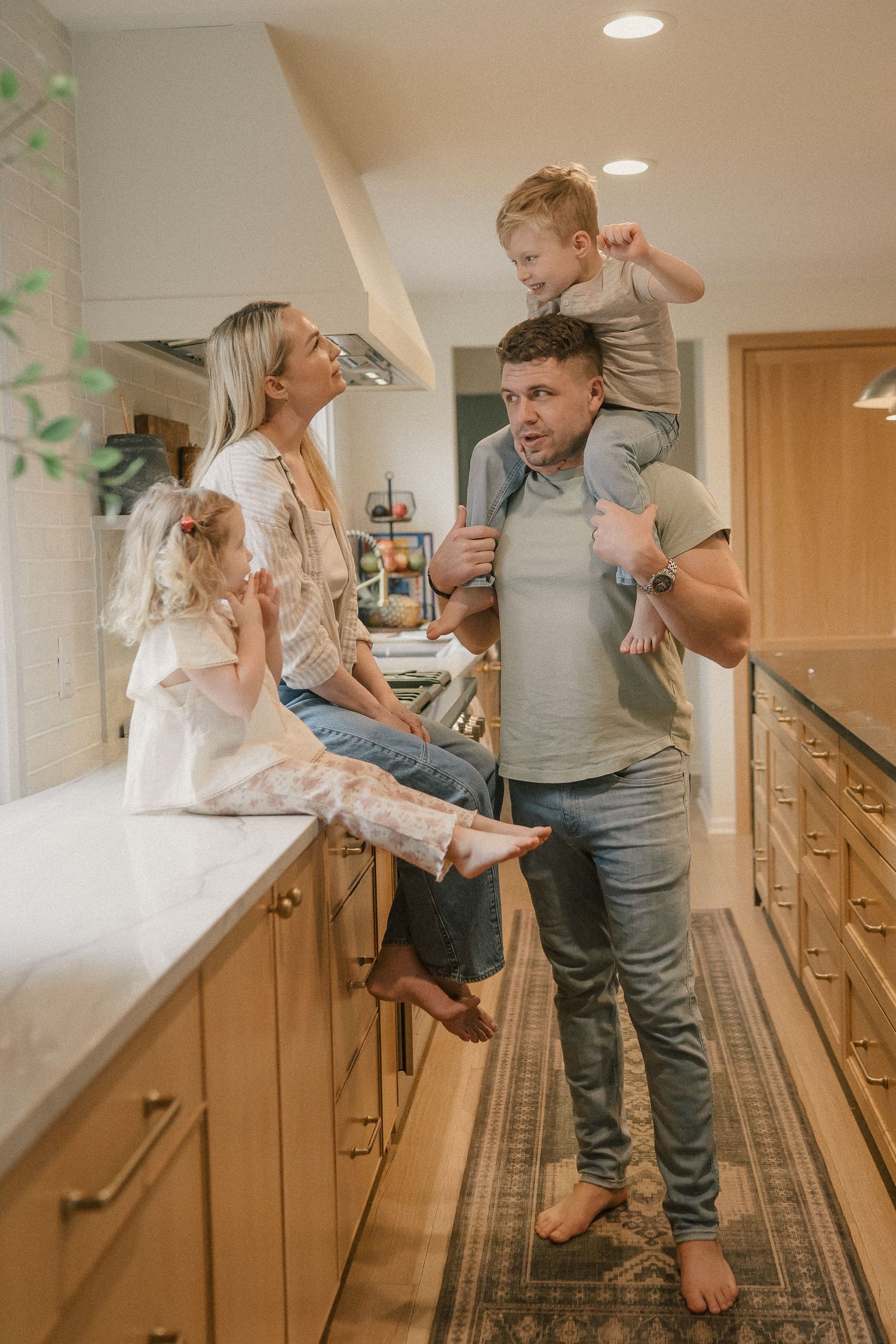 Family of four enjoys playful moment in modern kitchen, dad giving son piggyback ride while mom sits on counter with daughter.