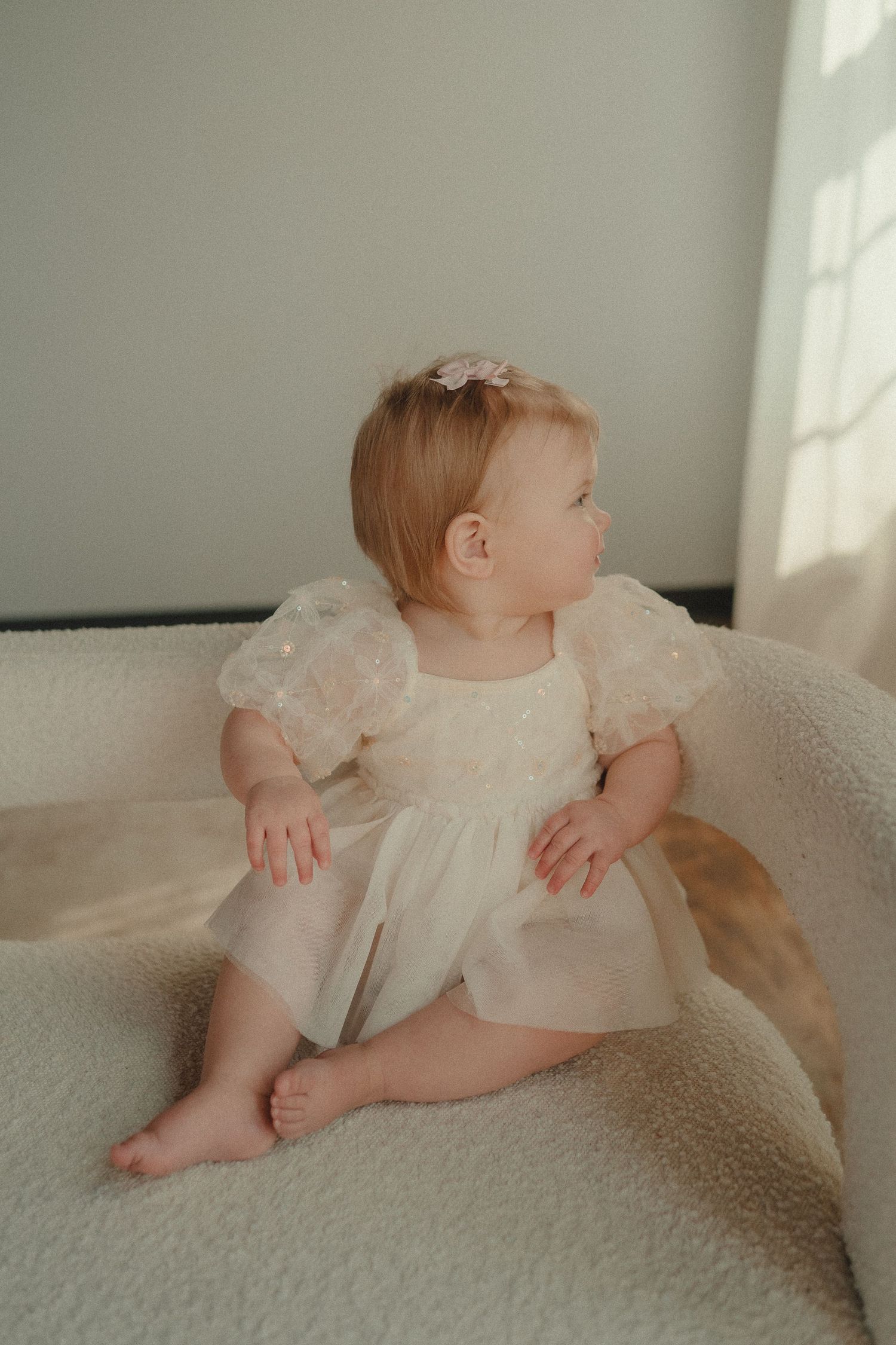 Adorable baby girl in white tulle dress with pink bow sitting on a cream textured chair.