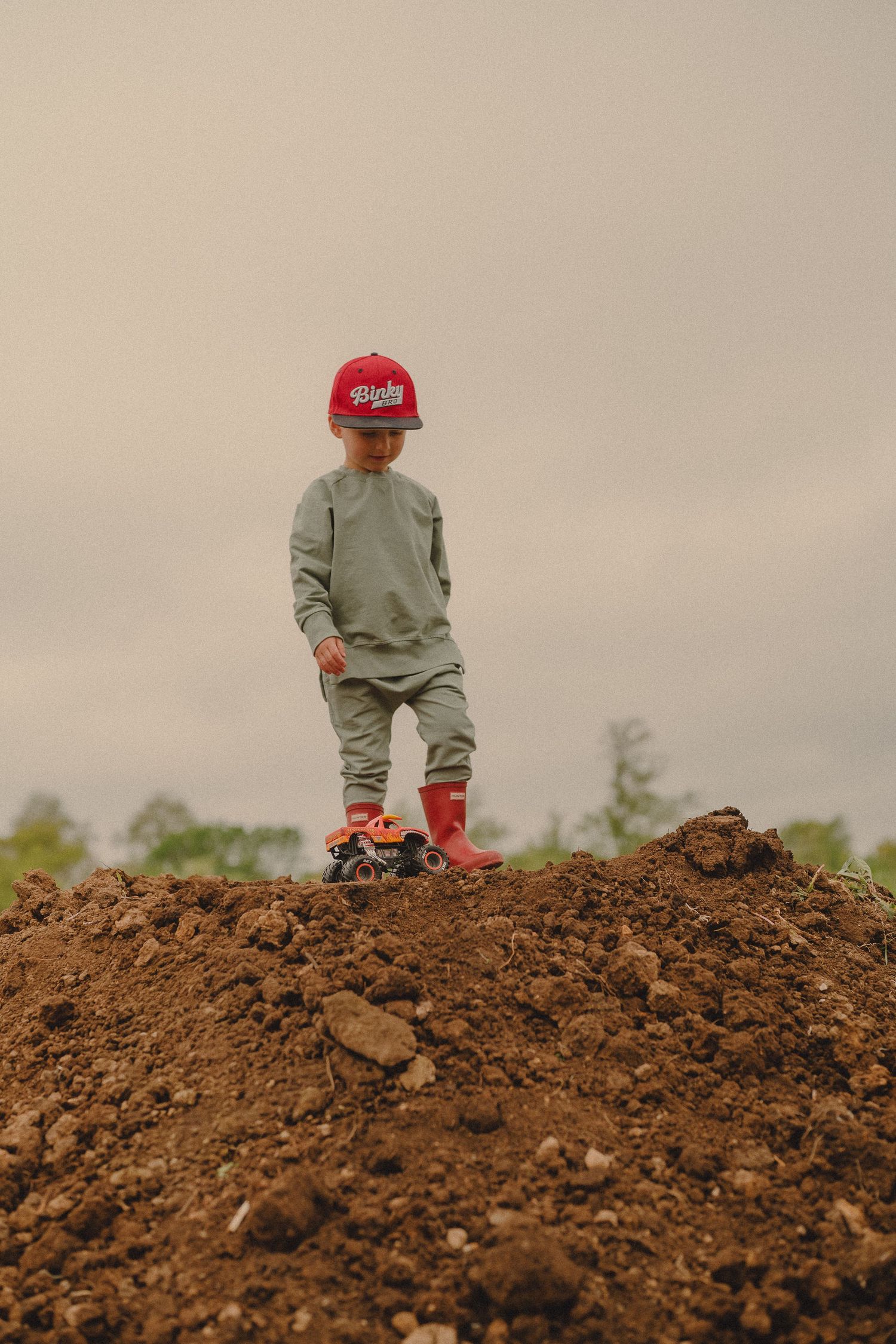 Young boy in red cap and boots standing triumphantly on a dirt mound outdoors.