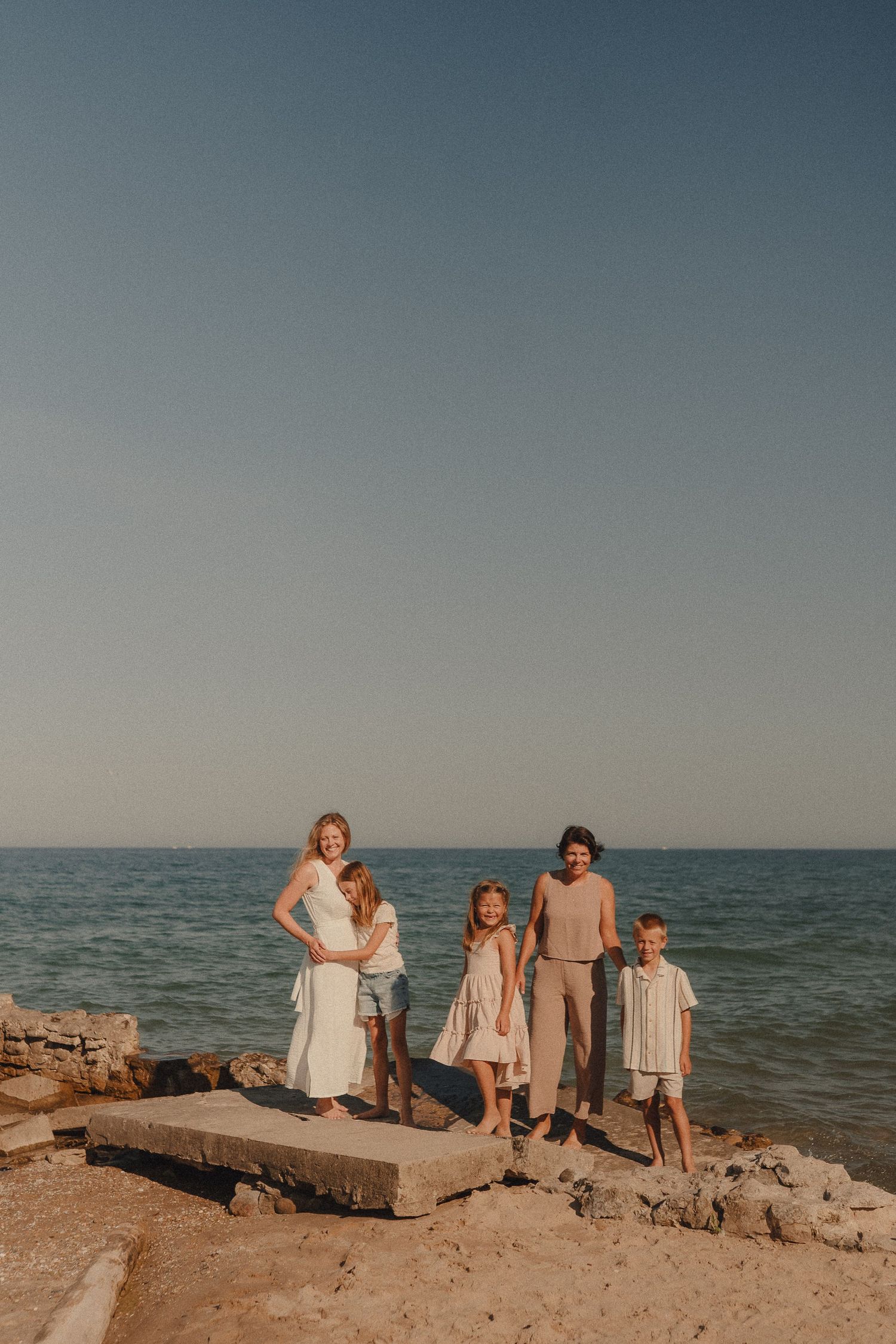 Family of five posing on rocky shoreline with calm blue ocean and clear sky backdrop on a sunny day.
